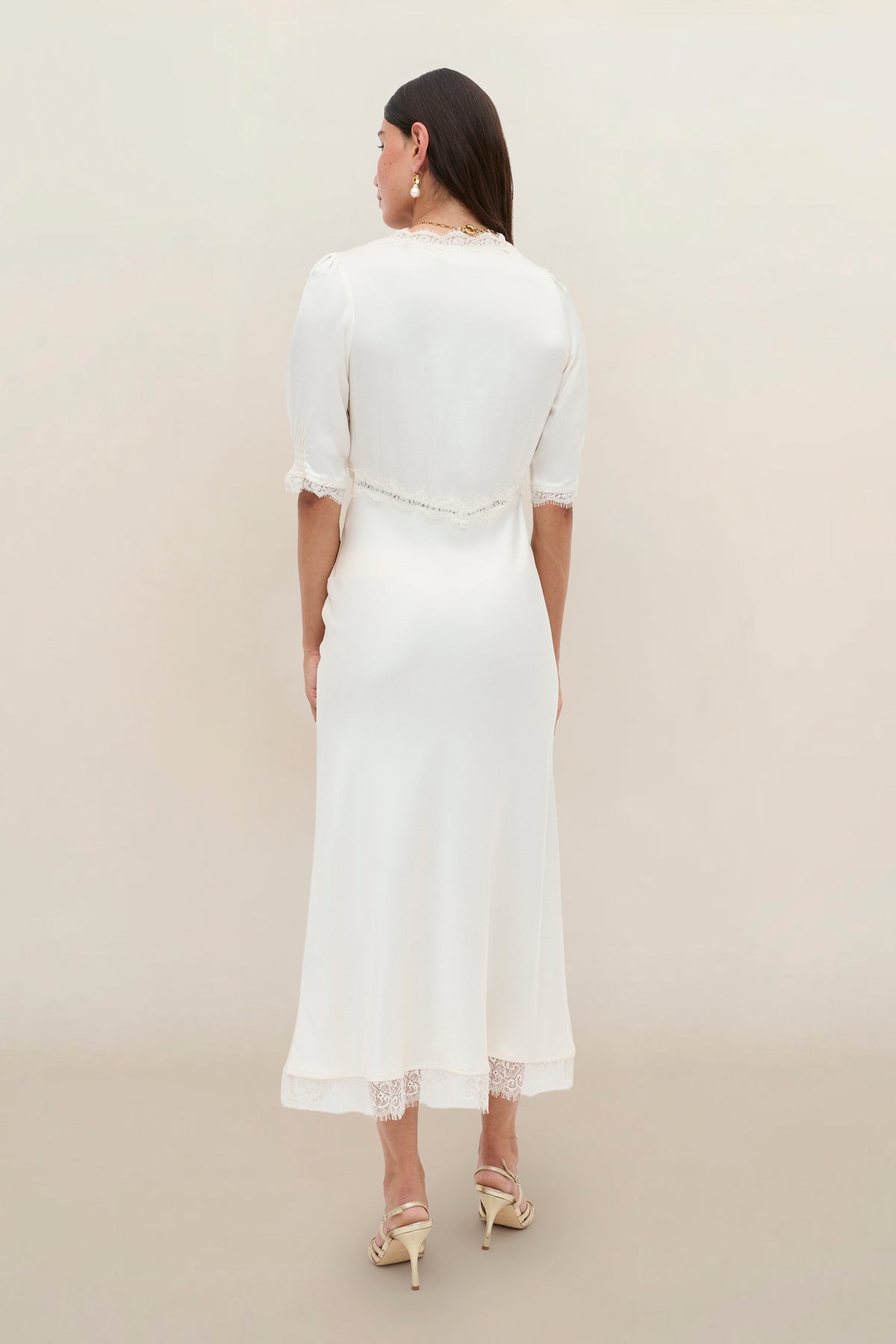 A woman with long dark hair, heeled sandals, and earrings stands facing away against a light background, wearing the Annina - Pearl dress featuring vintage-inspired lace trim and short sleeves.