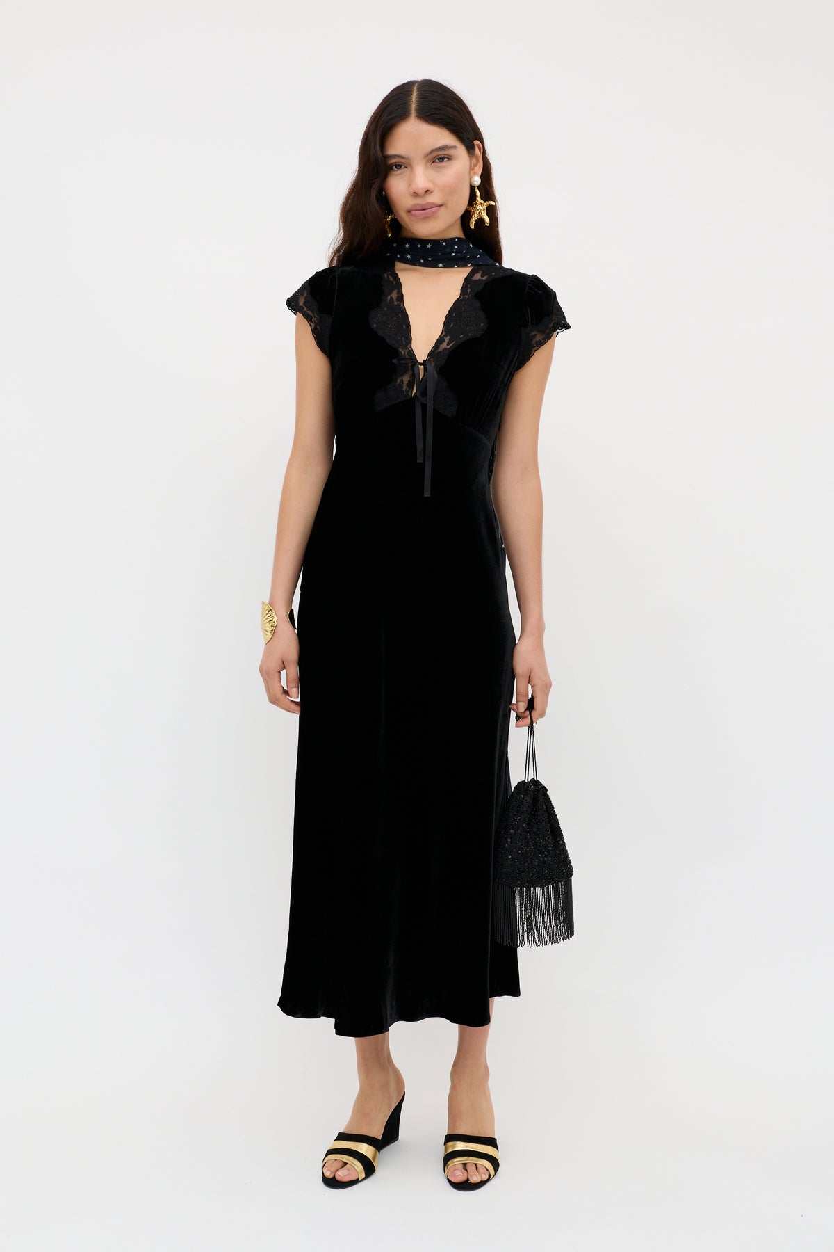 A woman in the Amari - Black velvet bias cut dress with lace, a matching scarf, black heeled sandals, gold earrings, and a small black beaded fringe handbag stands against a plain white background.