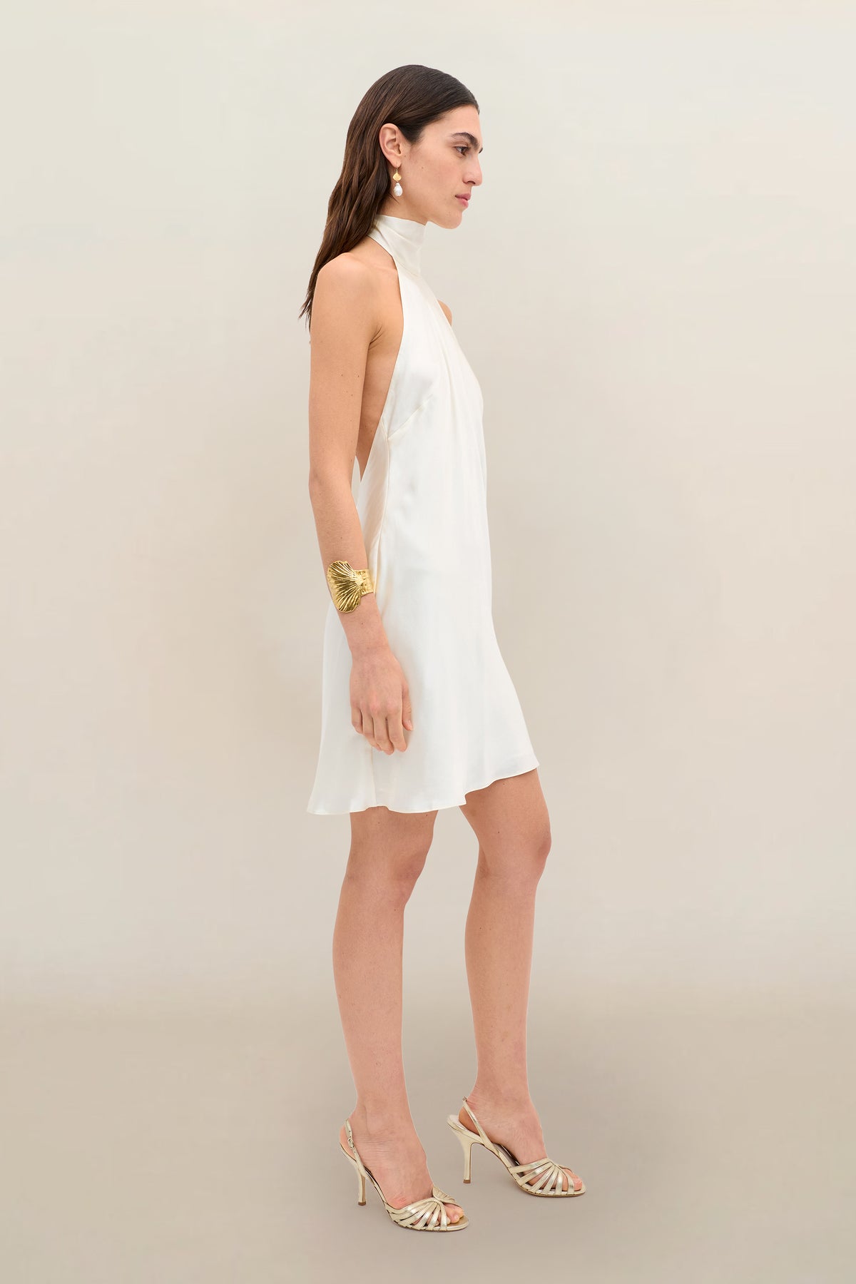 Wearing the Capucine - Champagne, a woman stands sideways against a plain beige background. The sleeveless, halter-neck party dress in lustrous champagne silk-satin features an open back, paired with heeled sandals. She completes her look with a large bracelet and drop earrings.