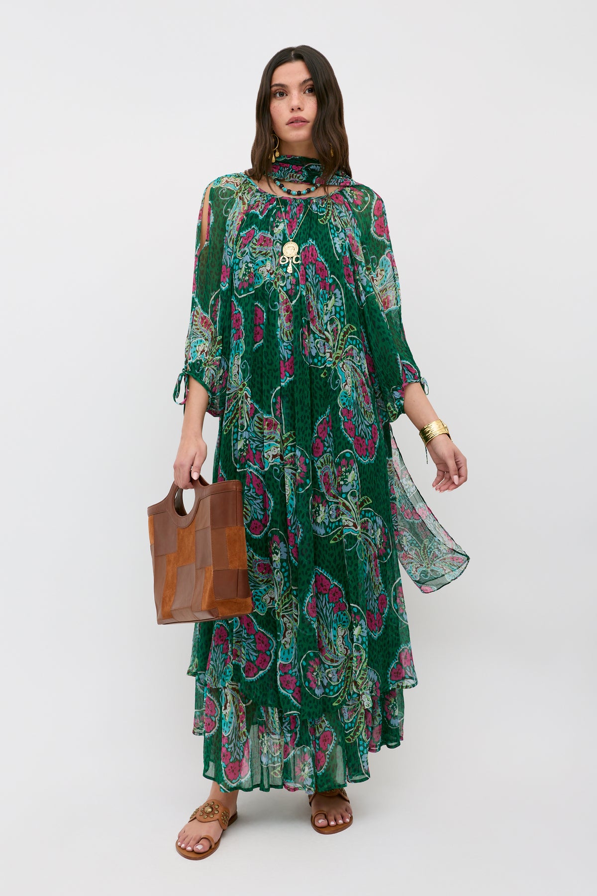 A woman stands against a plain background wearing the Hayden - Butterfly Bloom Green maxi dress with pink and blue patterns, sandals, gold jewelry, and holding a brown transparent handbag.