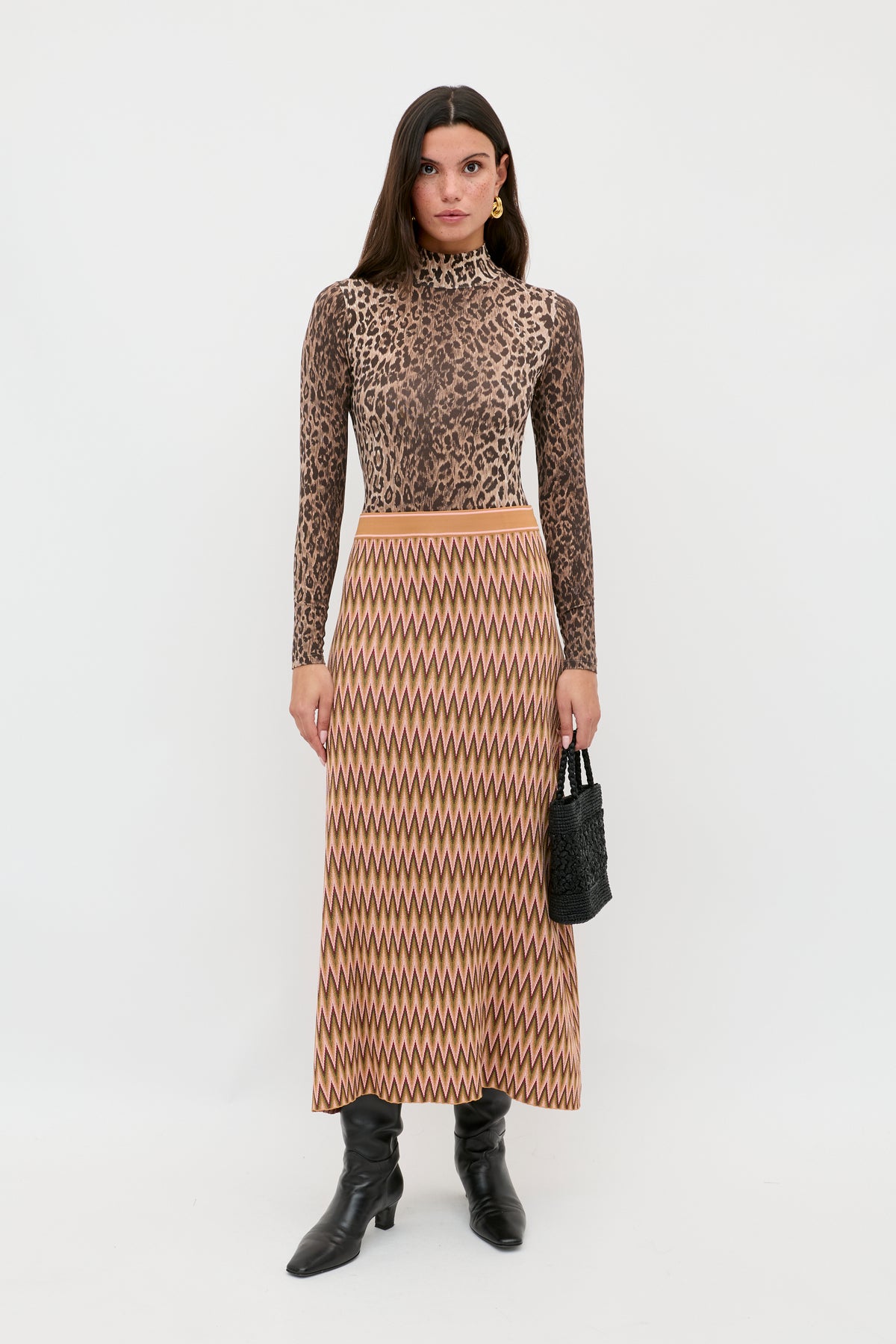 A woman stands against a white background wearing the Brandy - Maeve Jacquard Multi skirt with a leopard print turtleneck, black boots, and holding a small black textured handbag.