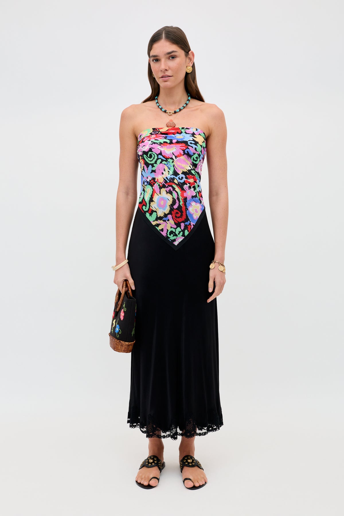 A woman wears the Crystal - Black dress, which features a colorful floral bodice and a flowing black satin midi skirt with delicate lace trim. She accessorizes with sandals, a necklace, a bracelet, and a handbag while standing against a plain white background.