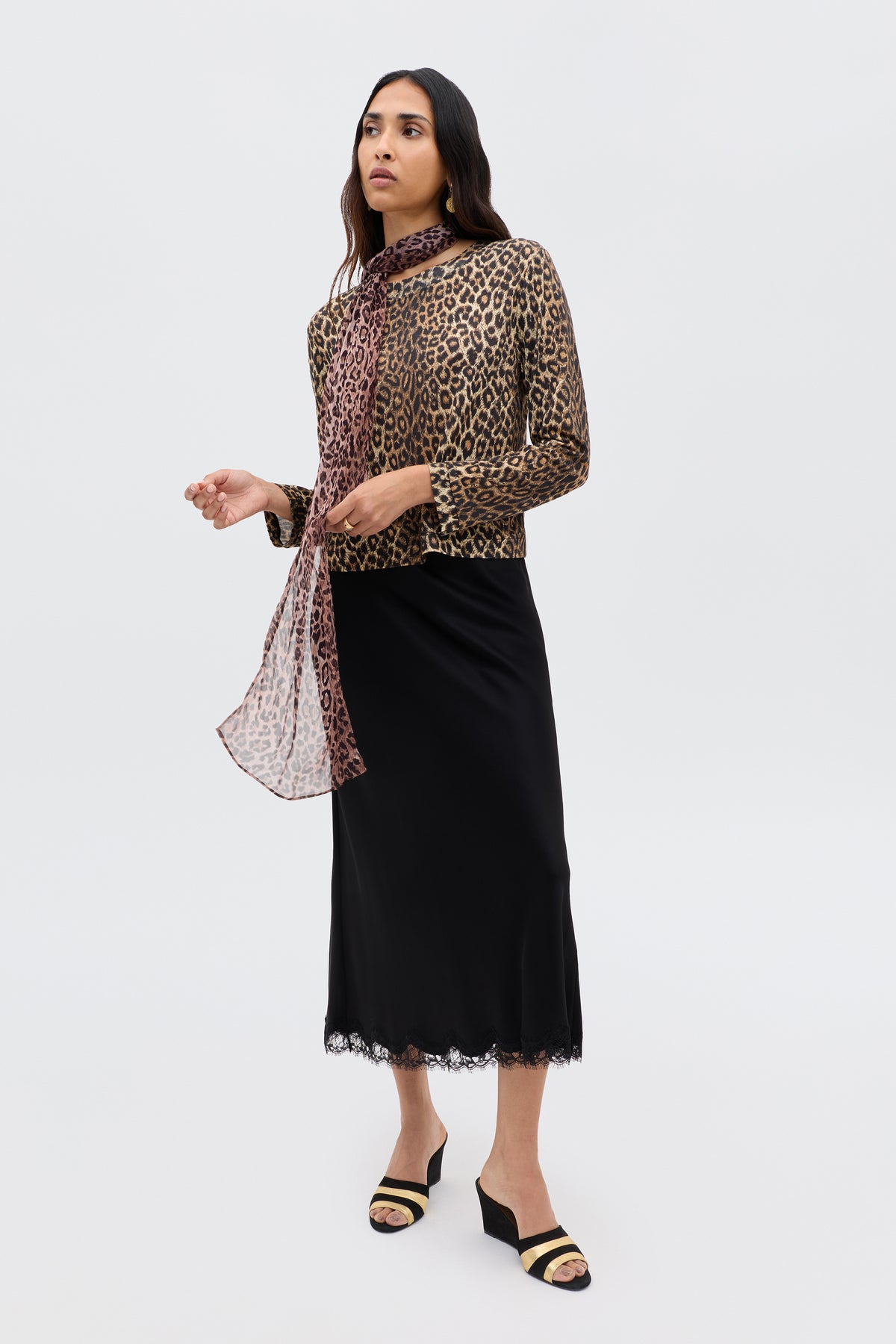 In Paris, a woman stands against a plain background, wearing the Stassie - Black sandals with a leopard-print top, sheer matching scarf, and a black satin skirt featuring wavy lace trim.
