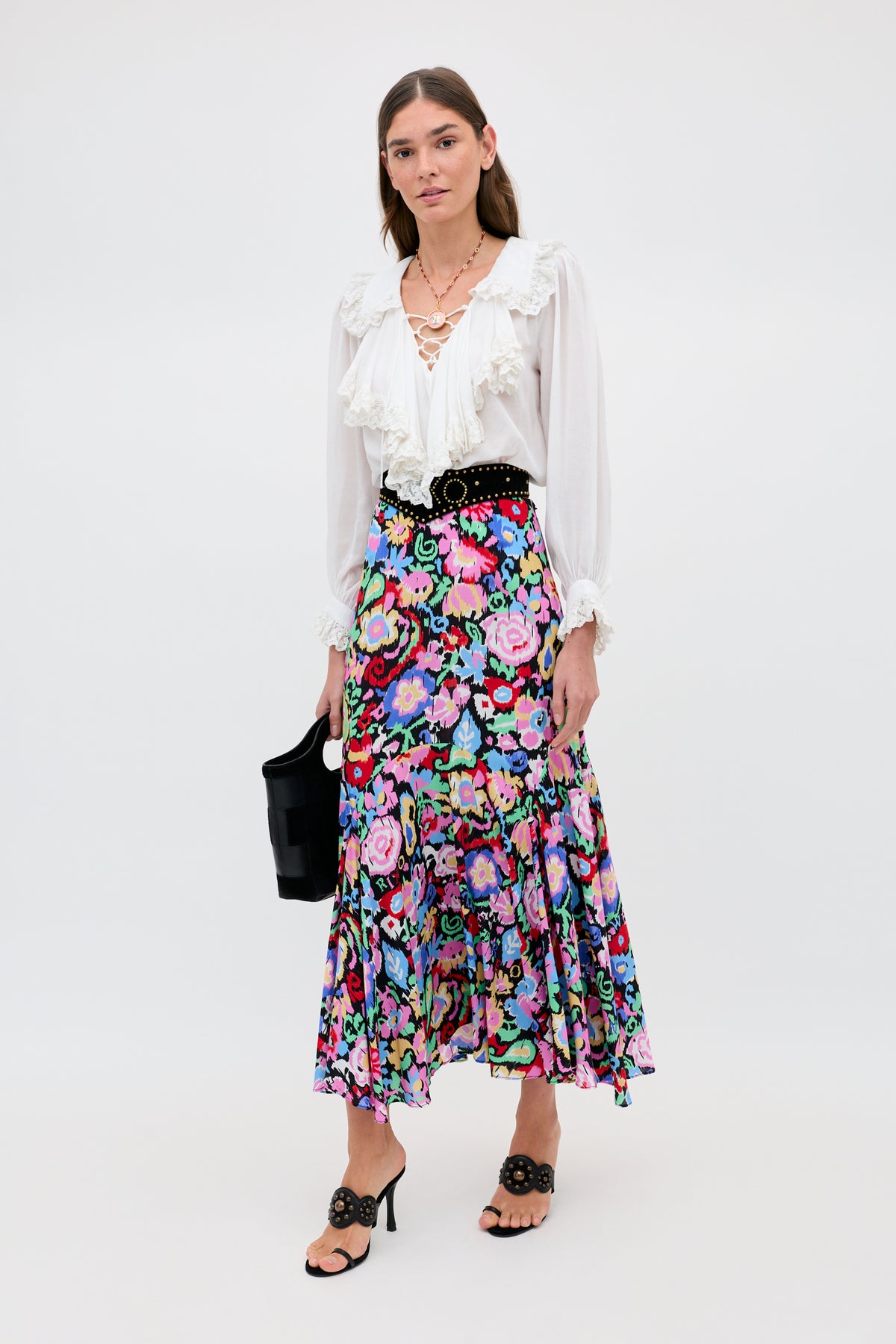 A woman poses against a plain background wearing the Libbie - Ikat Bloom Charcoal floral midi skirt, a white ruffled blouse, black sandals with a matching belt, layered necklaces, and carrying a black handbag.