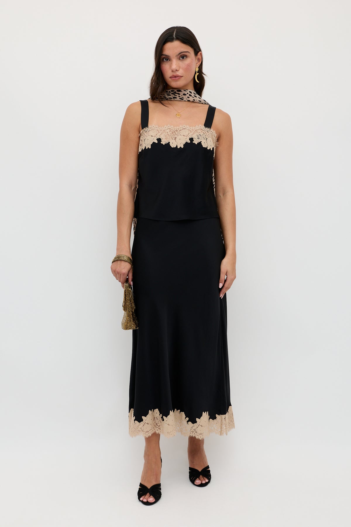A woman wearing the Matilde - Black sleeveless dress with beige lace trim at the neckline and hem, accessorized with a small gold bag, black heels, and a beige lace scarf, stands against a plain white background.