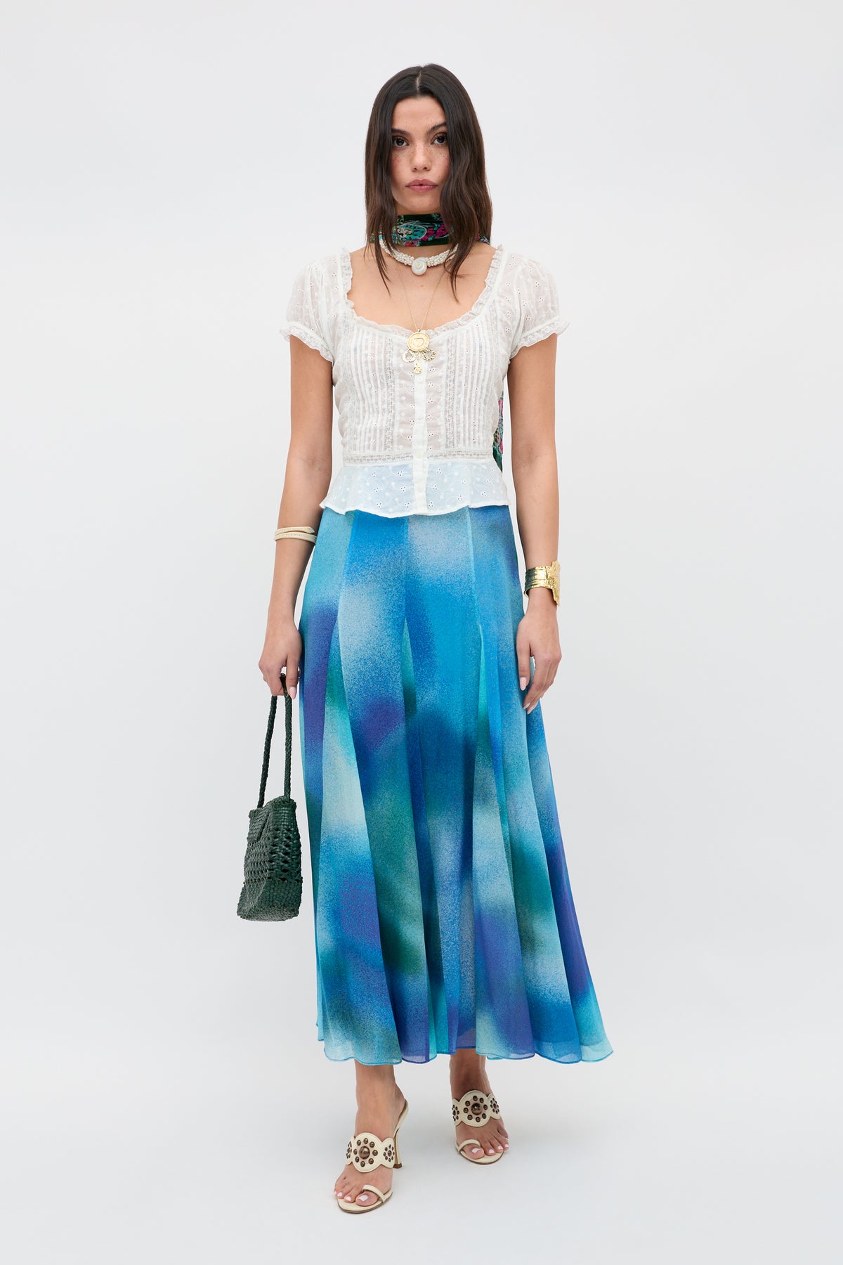 Woman wearing a white short-sleeve blouse, flowy Billy - Jessica Alexandra Blue ombre midi skirt crafted from Liberty fabric, white sandals, gold jewelry, a green patterned scarf around her neck, and holding a green textured handbag—perfect for effortless spring/summer dressing.