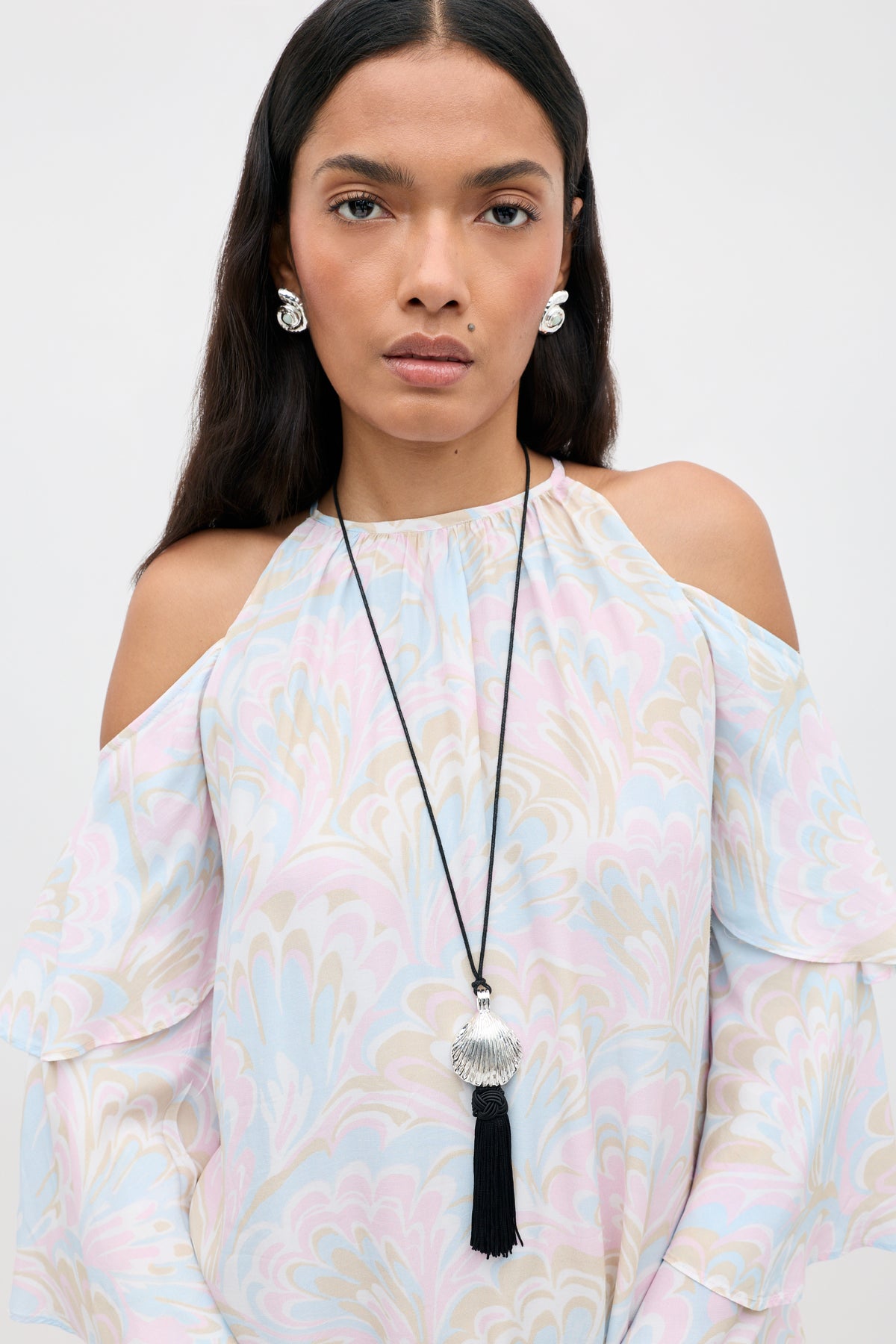 A woman wearing a pastel patterned off-shoulder blouse, silver earrings, and the Liana - Silver long necklace featuring a silver shell pendant with a black tassel, stands against a plain light background.