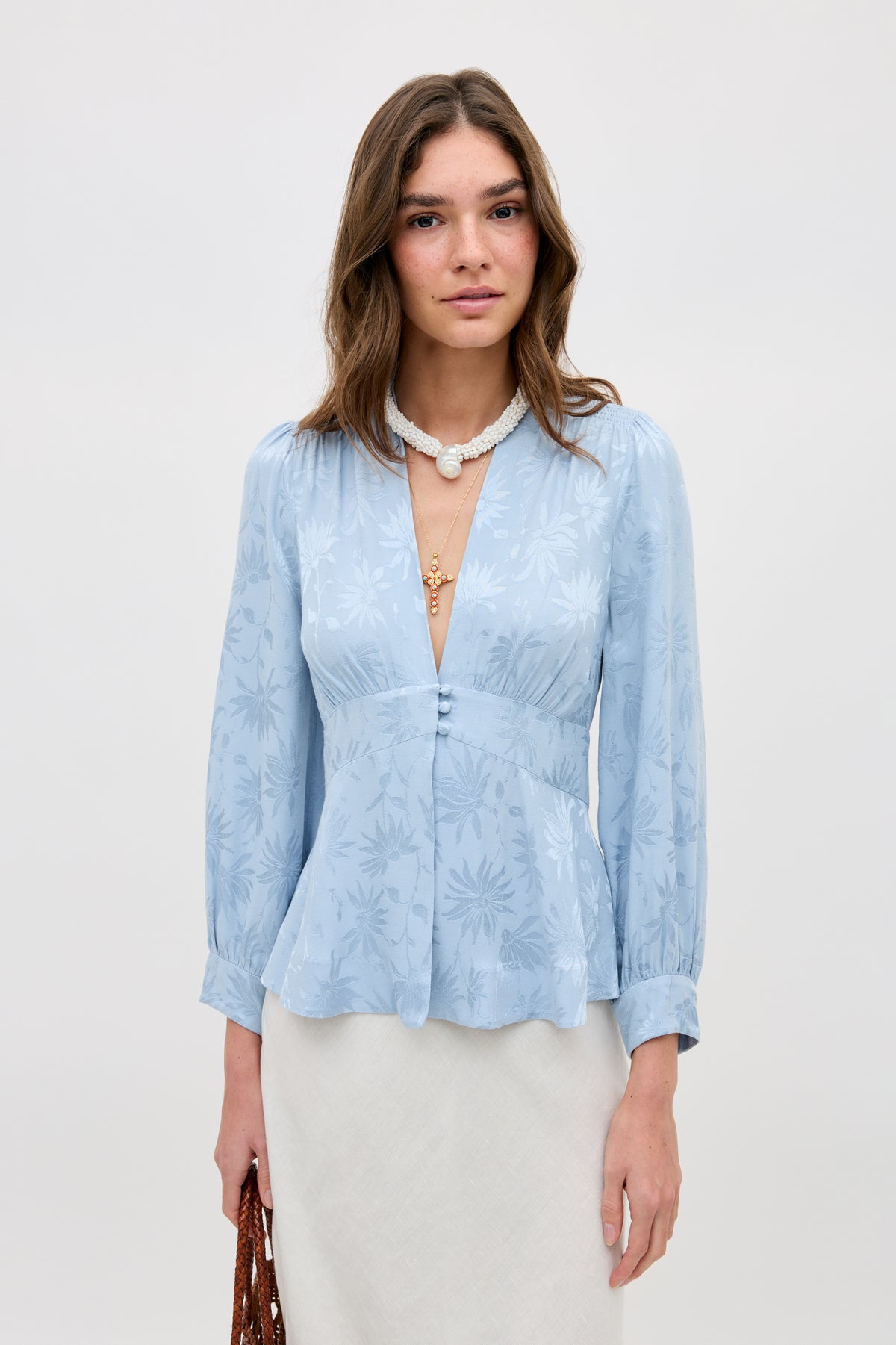 A woman stands against a plain background wearing the Lea - Daisy Jacquard Bluebell, a light blue, long-sleeve blouse with a floral pattern and deep V-neck. She holds a brown woven bag and accessorizes with a white necklace featuring a pendant.