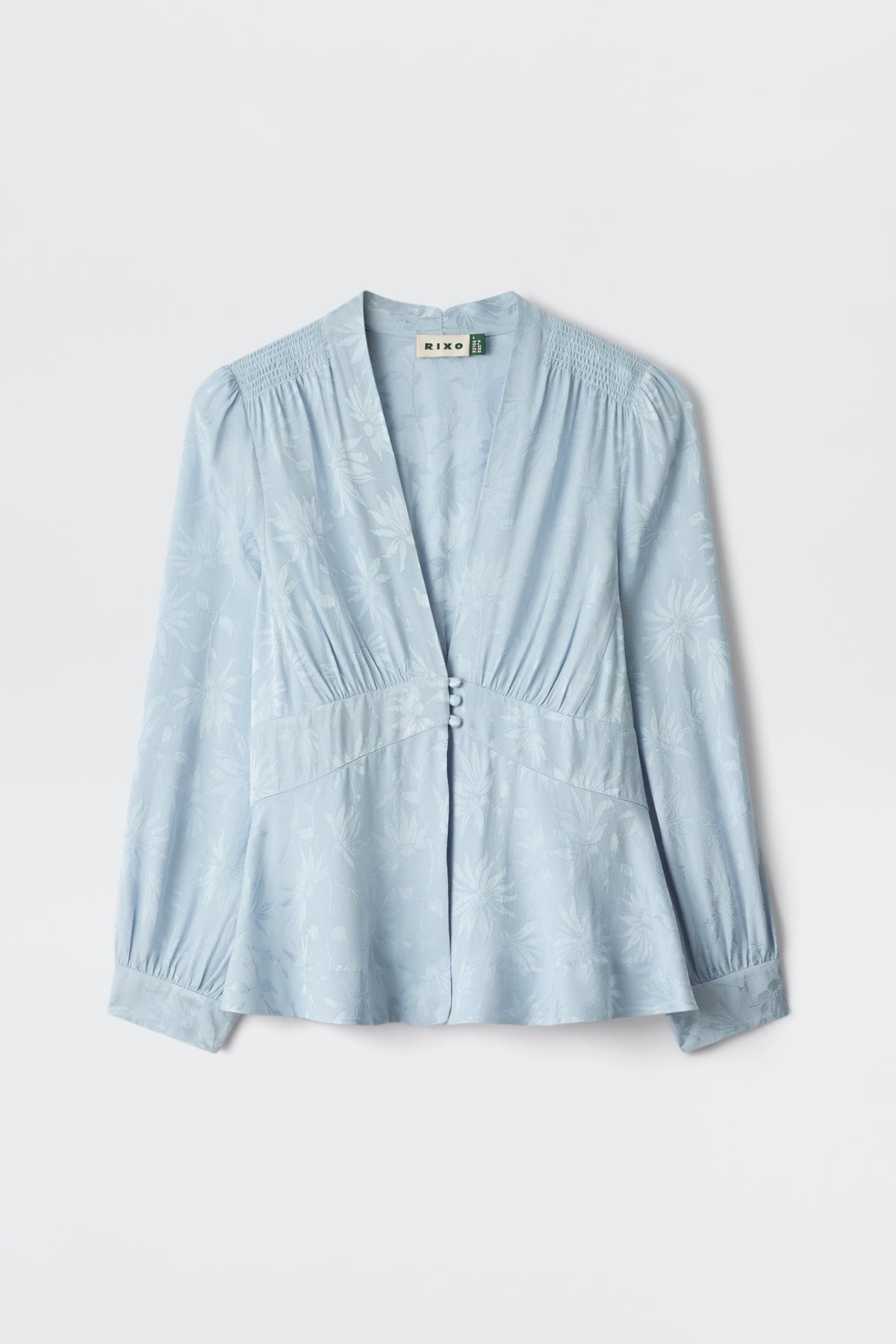 The Lea - Daisy Jacquard Bluebell is a light blue, long-sleeved blouse featuring a v-neckline, button front, subtle floral jacquard pattern, gathered shoulders, and a gently flared waist, shown against a plain white background.