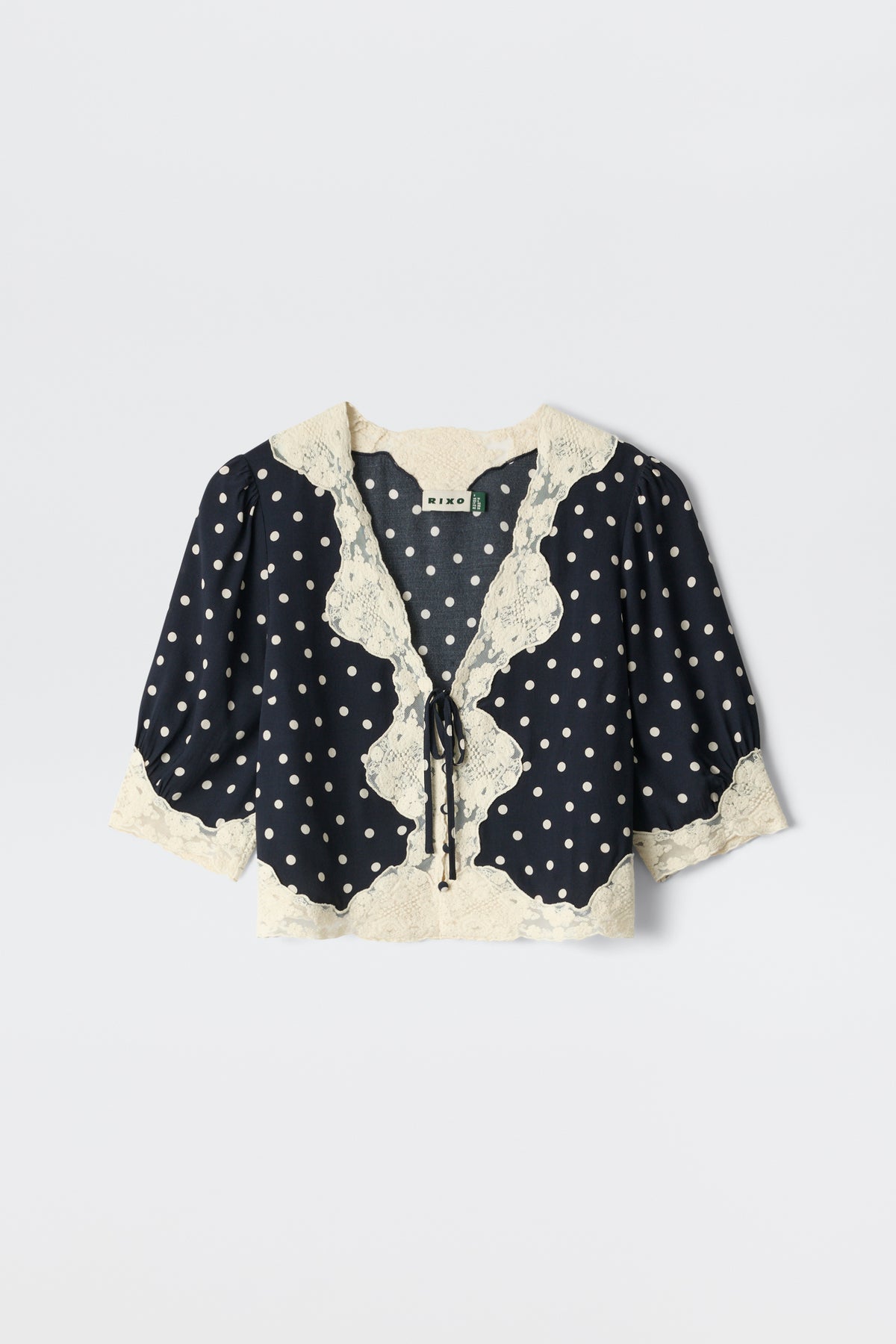 The Ursula - Layla Dot Navy is a navy blouse with white polka dots, puffed half sleeves, cream lace trim on the collar, front, and hem, plus a front tie closure.