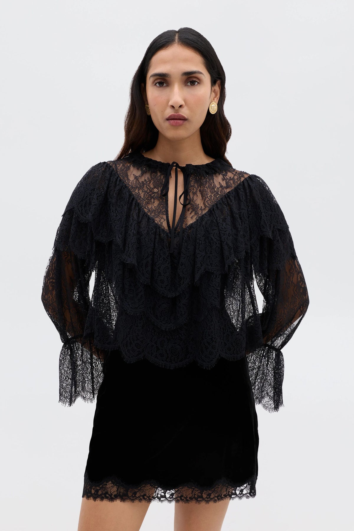 A woman with long dark hair wears the Faryn - Black, a sheer lace ruffle blouse with layered ruffles and long sleeves, paired with a black skirt. She stands against a plain light background, looking directly at the camera.