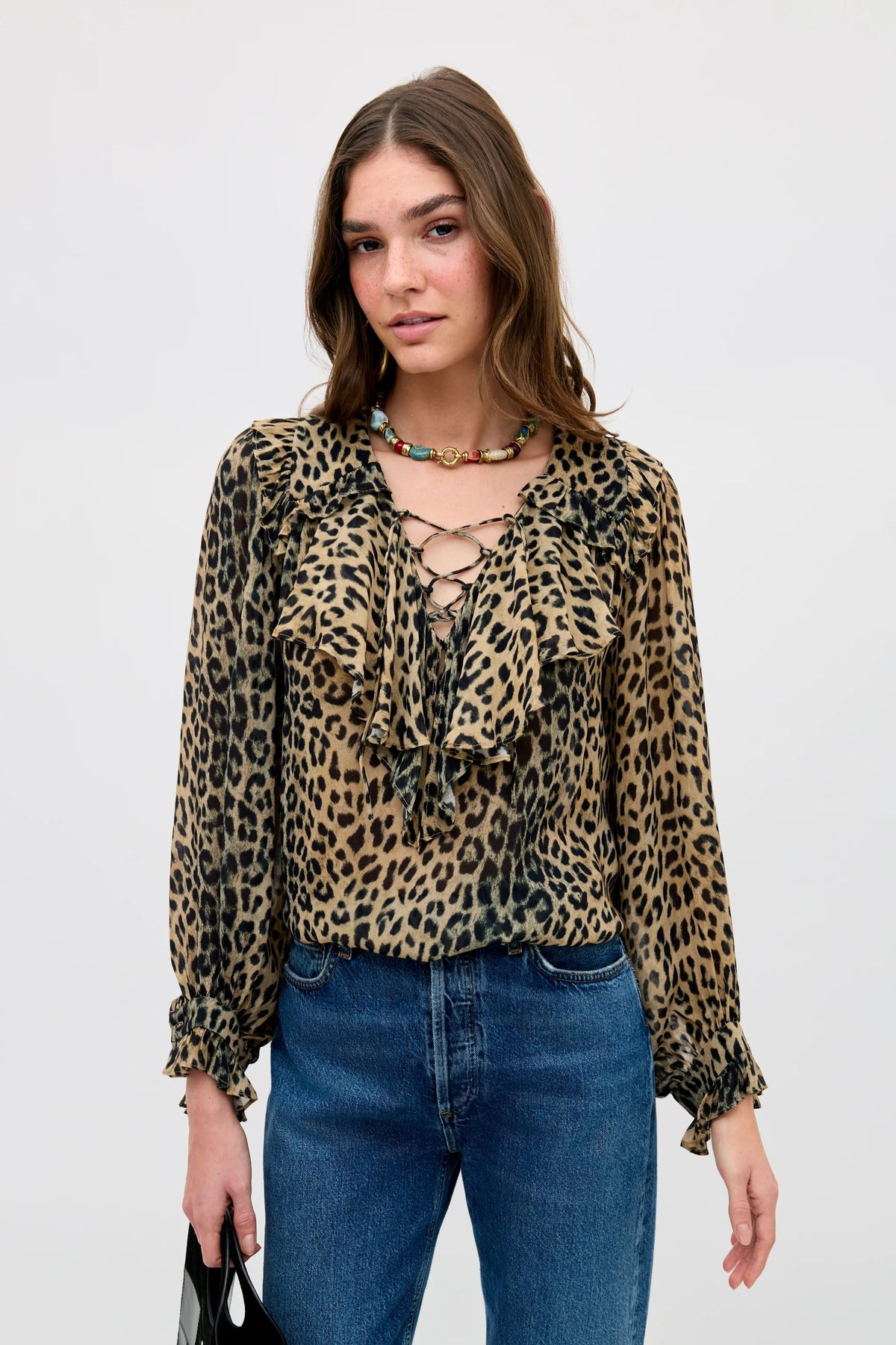 A woman stands against a plain light background wearing the Milia - Nina Leopard Brown blouse with ruffles and a lace-up front, paired with blue jeans and a beaded necklace. She holds a black bag.