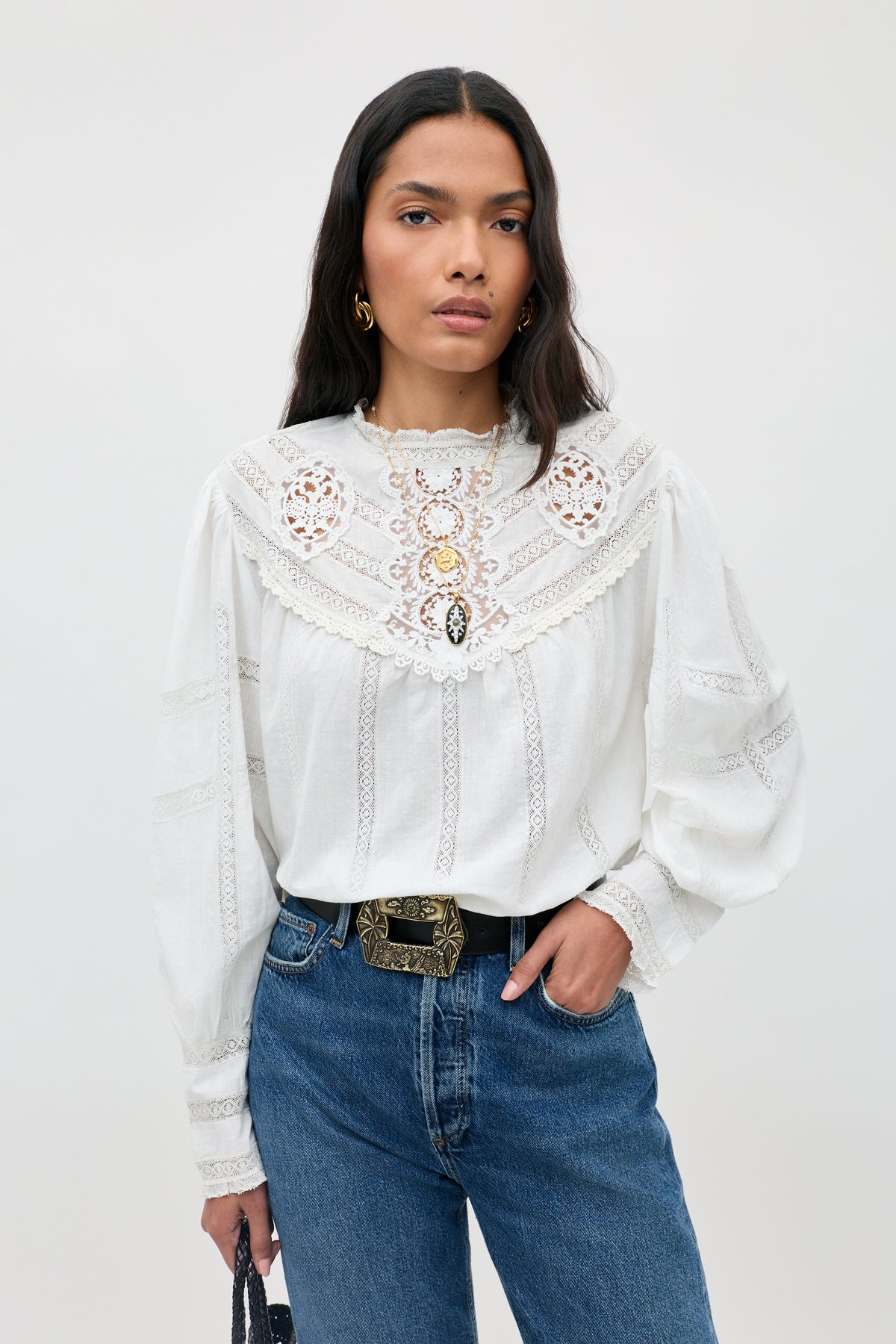 Wearing the Tamzin - Cream blouse, a woman with long dark hair stands against a light gray background, styled with blue jeans, an ornate belt, and gold earrings.