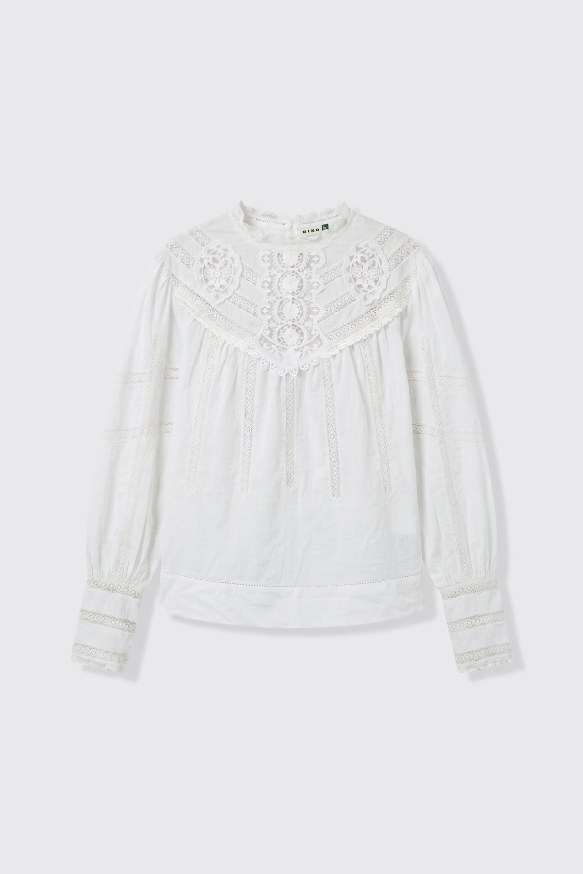 The Tamzin - Cream blouse features long sleeves and a high neckline, accentuated by lace details on the chest, sleeves, and cuffs. Its lightweight, textured fabric and intricate embroidery lend a vintage and elegant touch to the design.