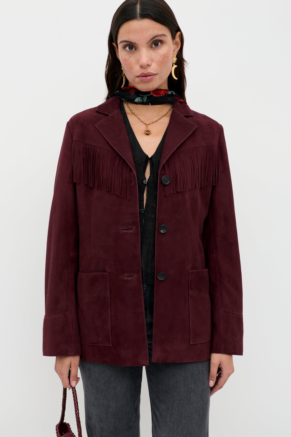 A woman wears the Teagan - Maroon suede fringe jacket, paired with a black shirt, dark jeans, layered necklaces, gold hoops, a floral scarf, and holds a small bag against a plain background.