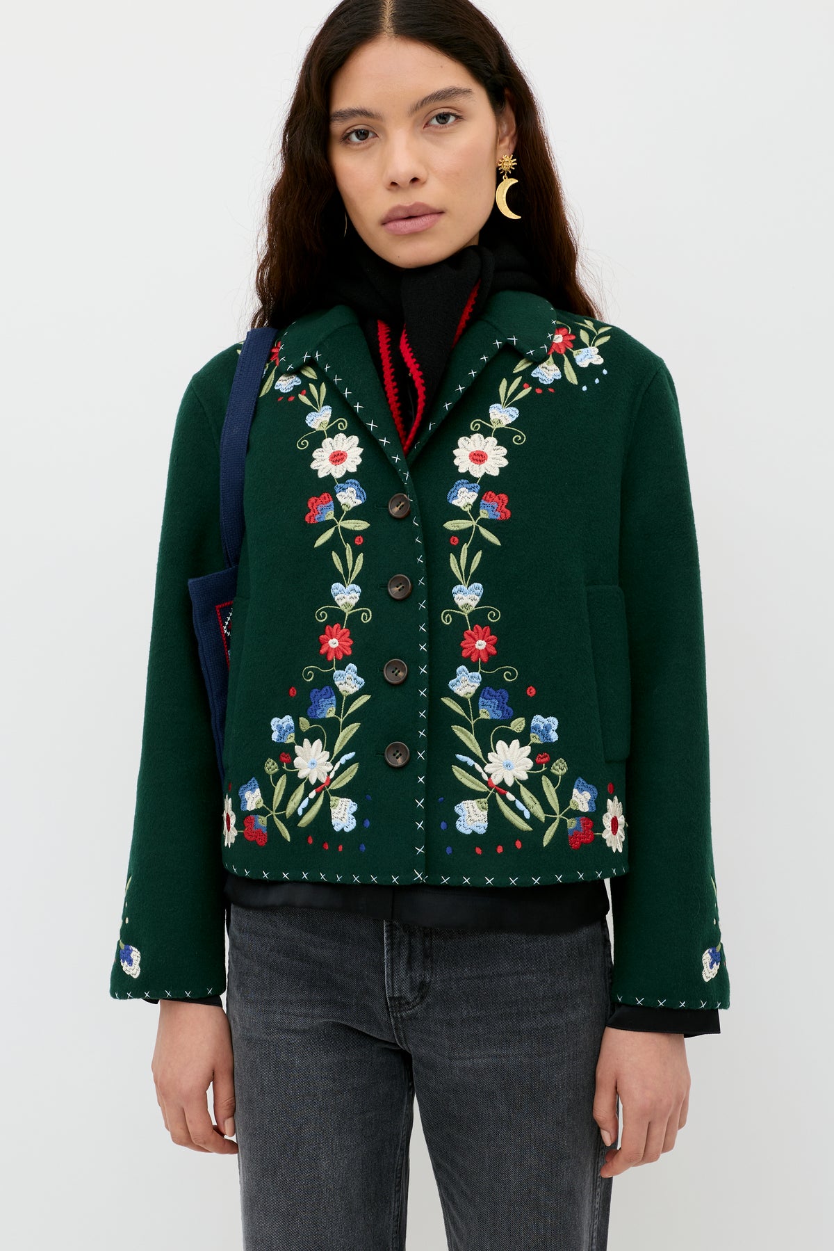 A woman with long dark hair wears the Jude - Bliss Embroidery Green jacket, featuring colorful embroidery, along with a black and red sweater, dark jeans, and crescent moon earrings, standing against a plain white background.
