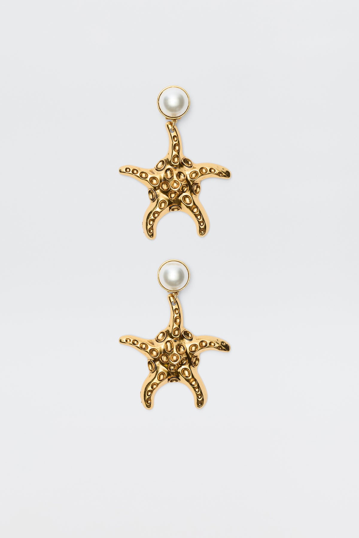 Zeina - Gold earrings featuring starfish shapes with textured surfaces and a round pearl at the top, shown against a plain white background.