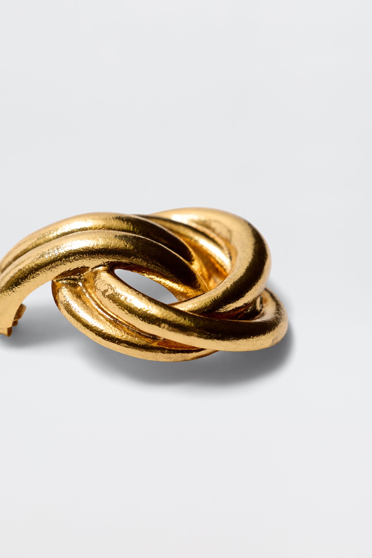 The Nolana - Gold ring is a shiny gold-plated brass piece with two thick, intertwined loops in a knot-like design, set against a plain light gray background.