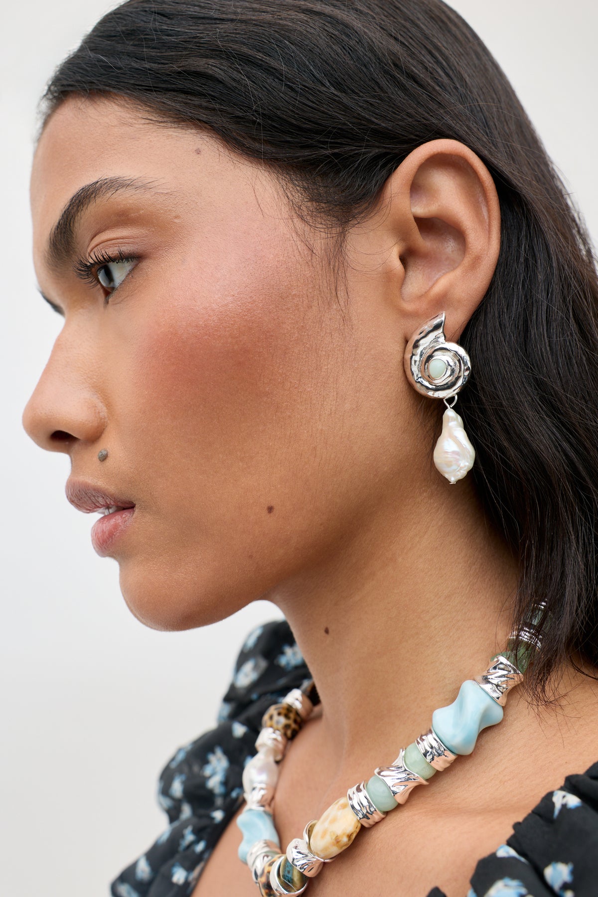 A person with long dark hair is wearing the Cali - Silver earring, featuring a pearl drop, along with a chunky necklace adorned with blue and beige stones. Their warm-toned skin complements their black floral top.