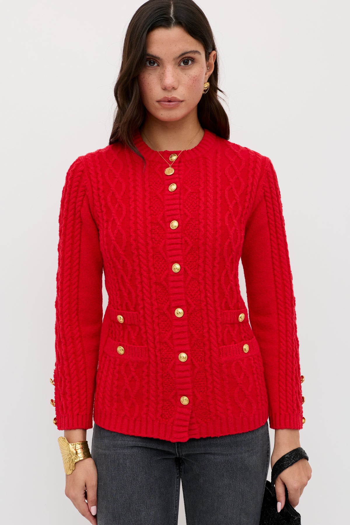 A woman with dark wavy hair wears the Maurya - Red cable-knit cardigan with gold buttons and front pockets, styled with dark gray jeans and gold jewelry against a plain white background.