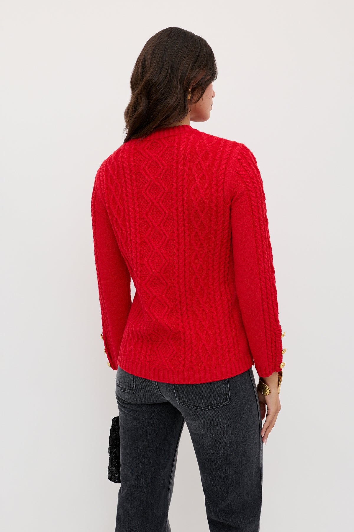 A woman with wavy brown hair stands with her back to the camera, wearing the Maurya - Red cable-knit sweater and black jeans. She holds a small black bag in her left hand against a plain white background.