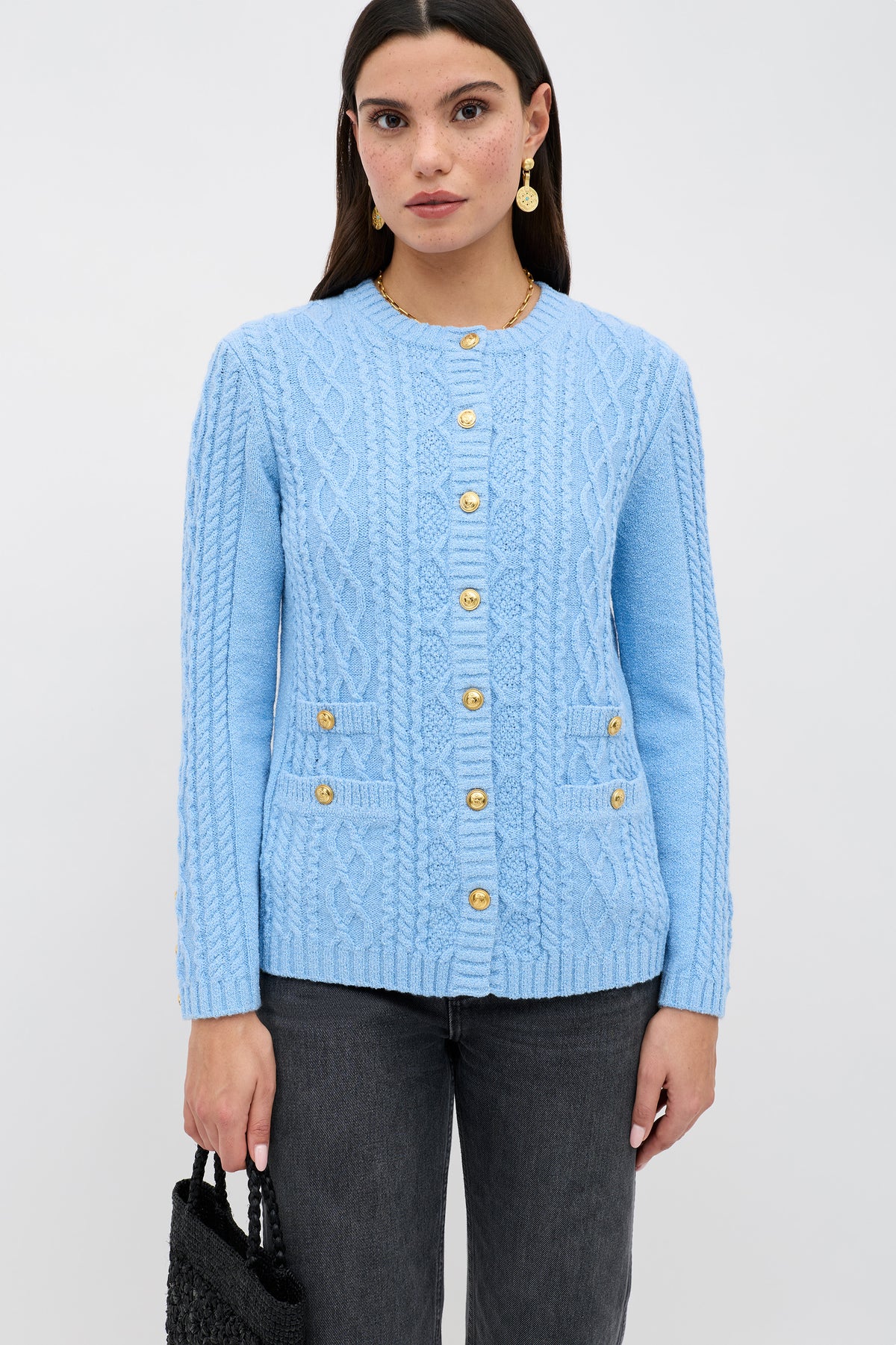 A woman wears the Maurya - Dusty Blue cable-knit cardigan with gold buttons and front pockets, styled with dark jeans, a black woven bag, and gold earrings, standing against a plain light background.