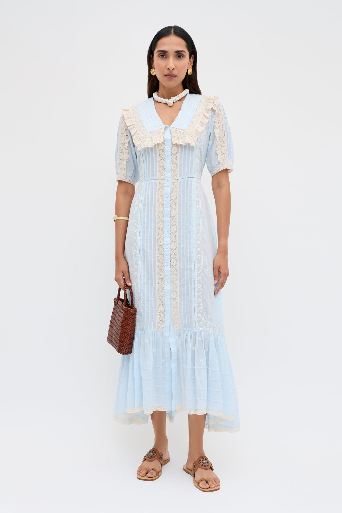A woman stands against a plain white background wearing the Edina - Mira Embroidery Powder Blue blouse with a ruffled hem, paired with a woven brown handbag, flat sandals, and simple jewelry.