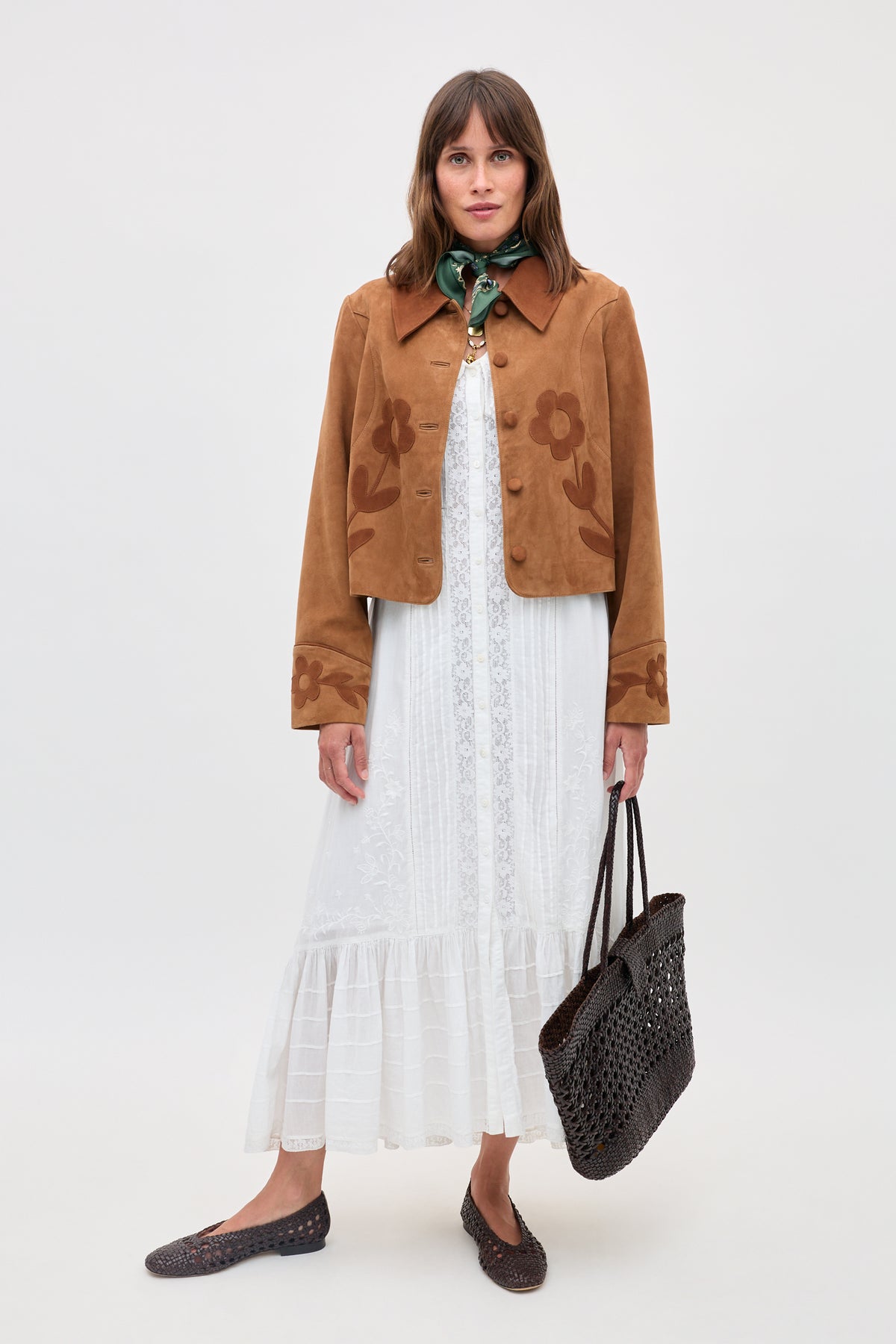 A woman models the Dionne - Retro Daisy tan suede jacket with daisy appliqué, styled over a white lace dress, green neck scarf, brown woven tote, and brown flats, standing against a plain light background.