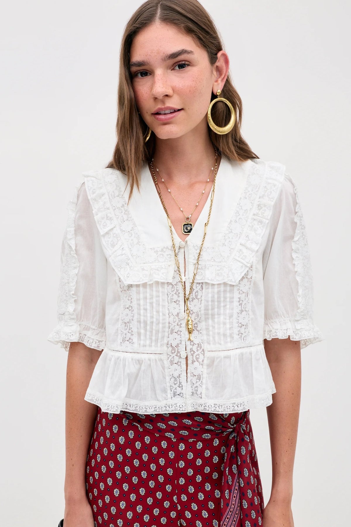 A woman wears the Ellery - White vintage-inspired sheer lace blouse with a ruffled collar and sleeves, paired with red patterned pants. She adds gold hoop earrings, layered necklaces, and carries a black bag.