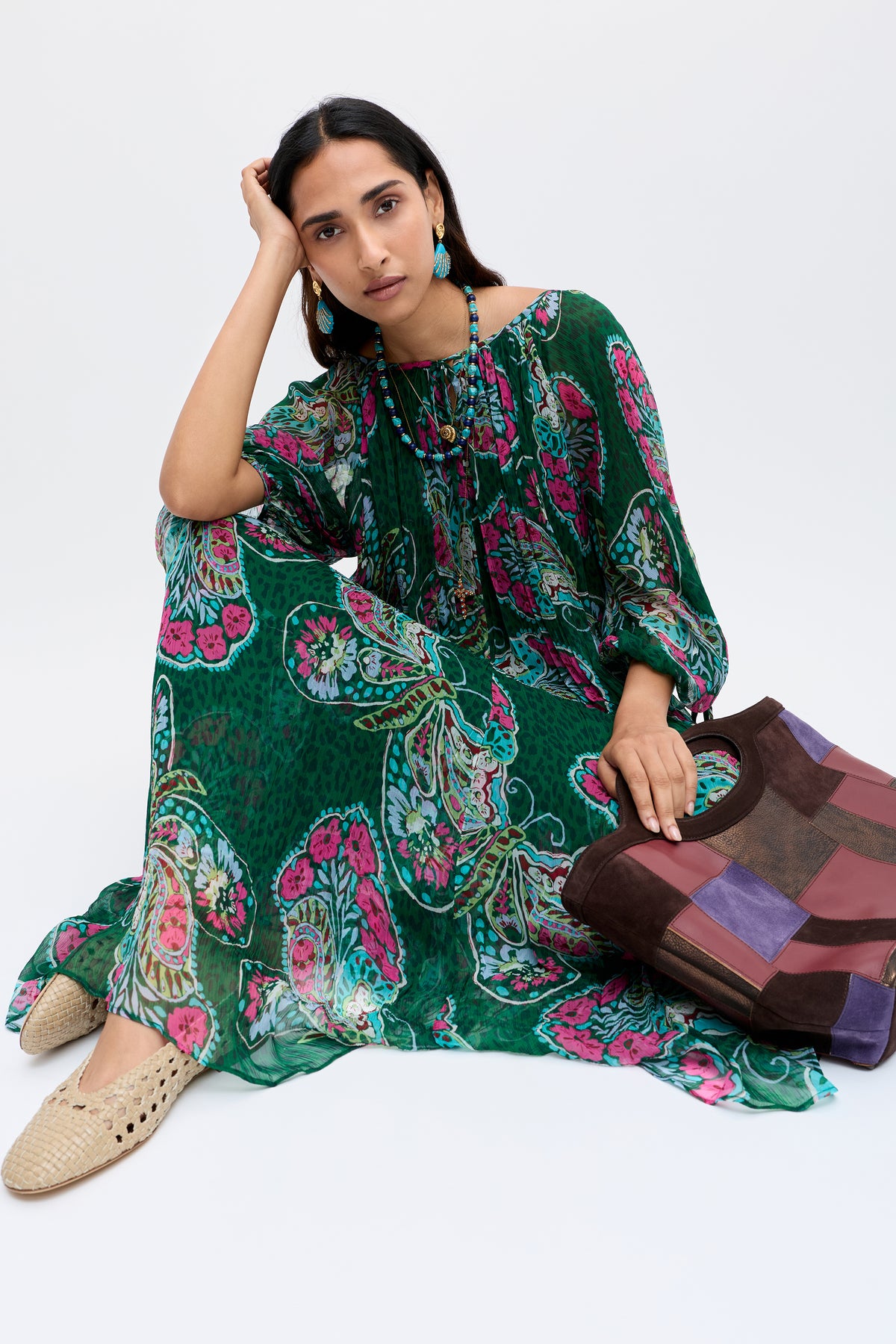 A woman in the Hayden - Butterfly Bloom Green dress sits on the floor, leaning her head on her hand. She accessorizes with woven shoes, blue earrings, colorful beaded necklaces, and a large patchwork bag against a white background.