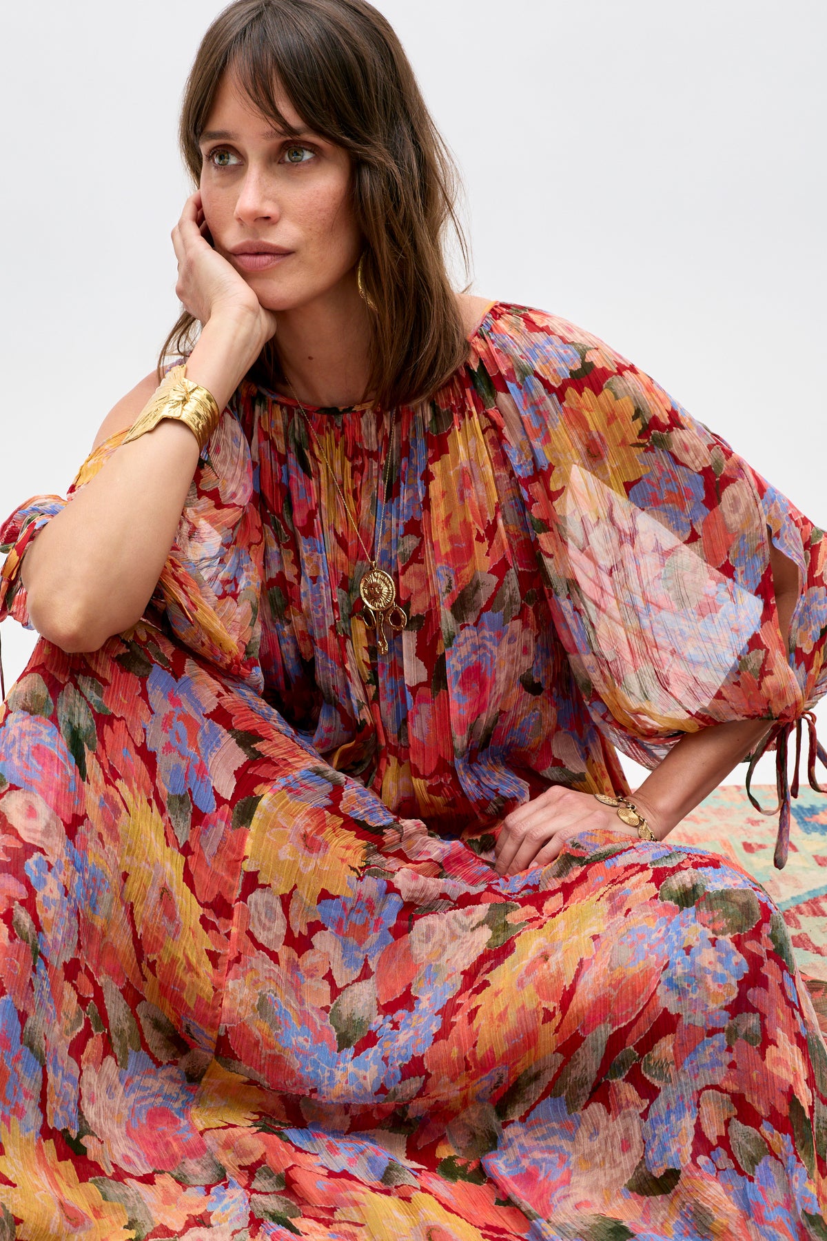 A woman wearing the Hayden - Chelsea Floral Red dress sits with her chin resting on her hand, adorned in gold jewelry and gazing thoughtfully to the side against a plain light background.