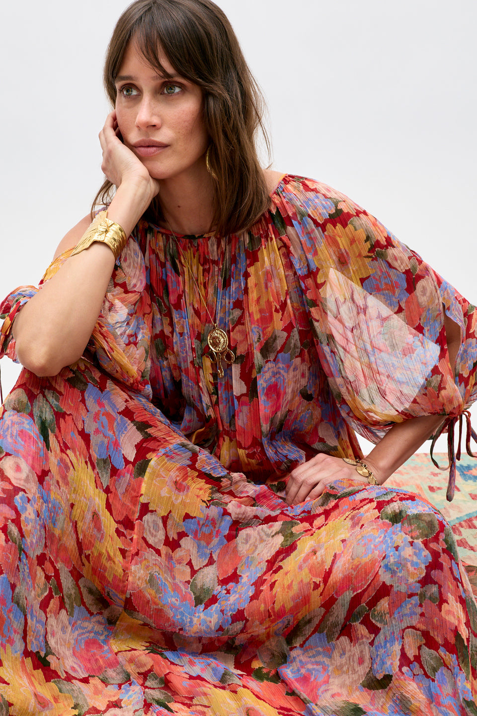 A woman wearing the Hayden - Chelsea Floral Red dress sits with her chin resting on her hand, adorned in gold jewelry and gazing thoughtfully to the side against a plain light background.