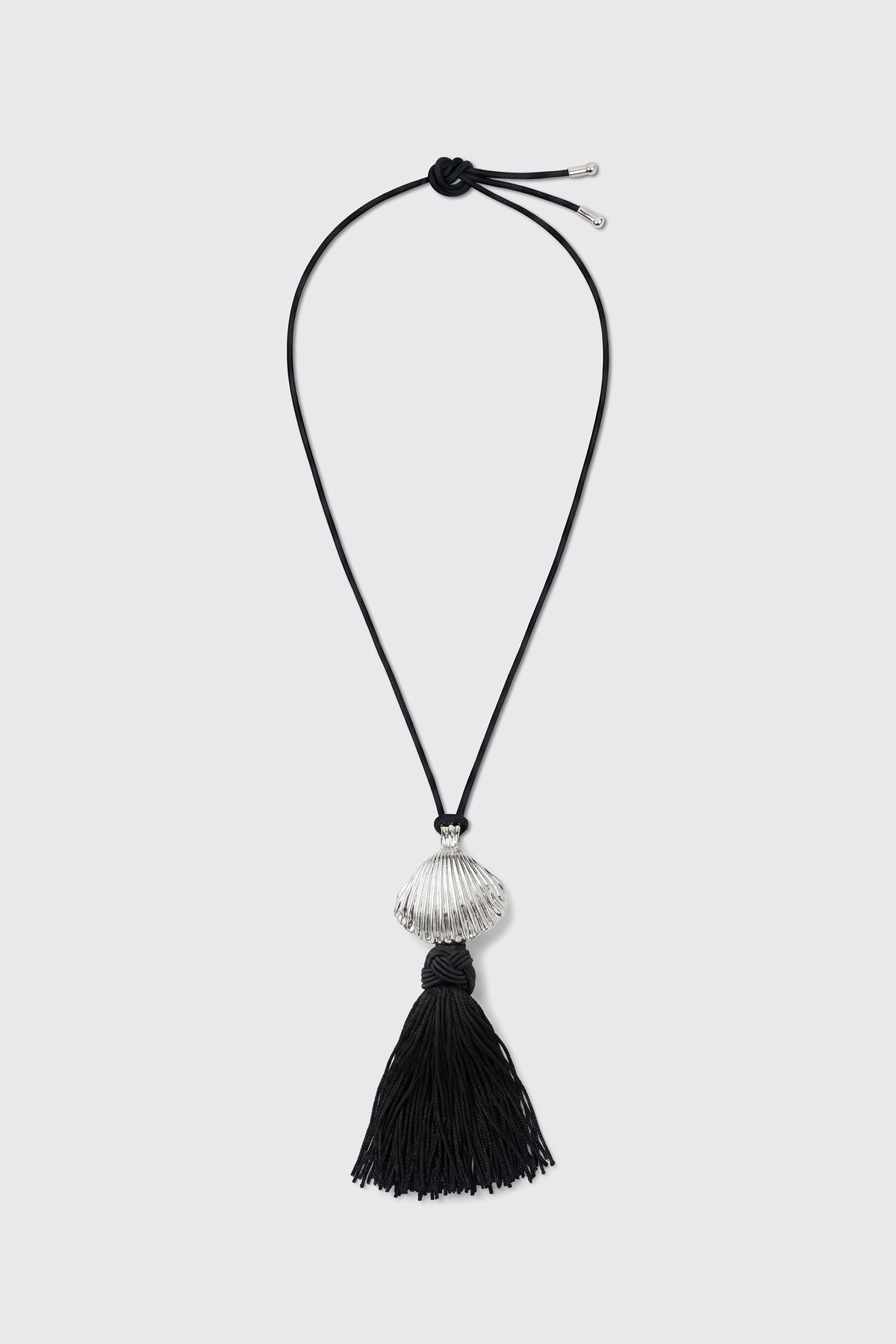 Liana - Silver: A black cord necklace featuring a silver shell-shaped bead and a large black tassel pendant, displayed on a plain light background.