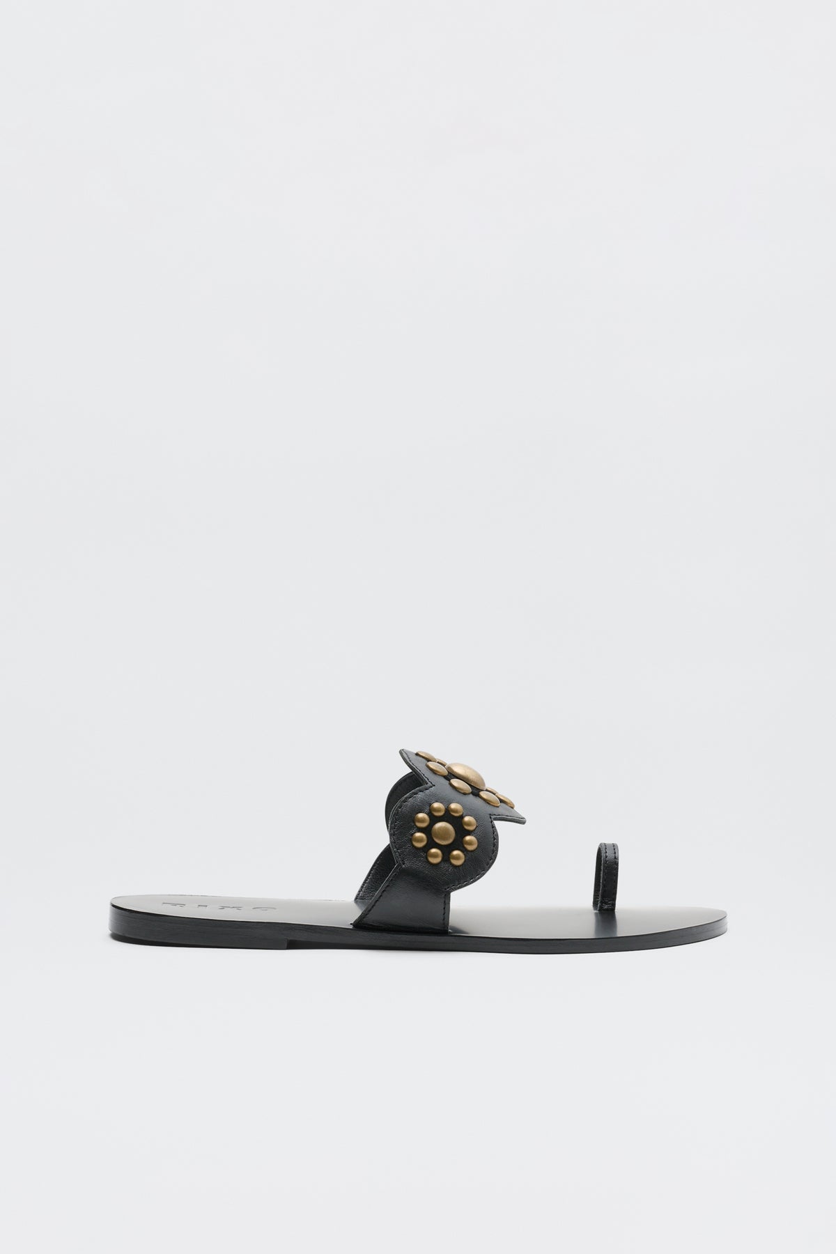 The Lucille - Black is a flat sandal featuring a toe loop and circular embellishment adorned with gold studs on the upper, showcased against a plain white background.