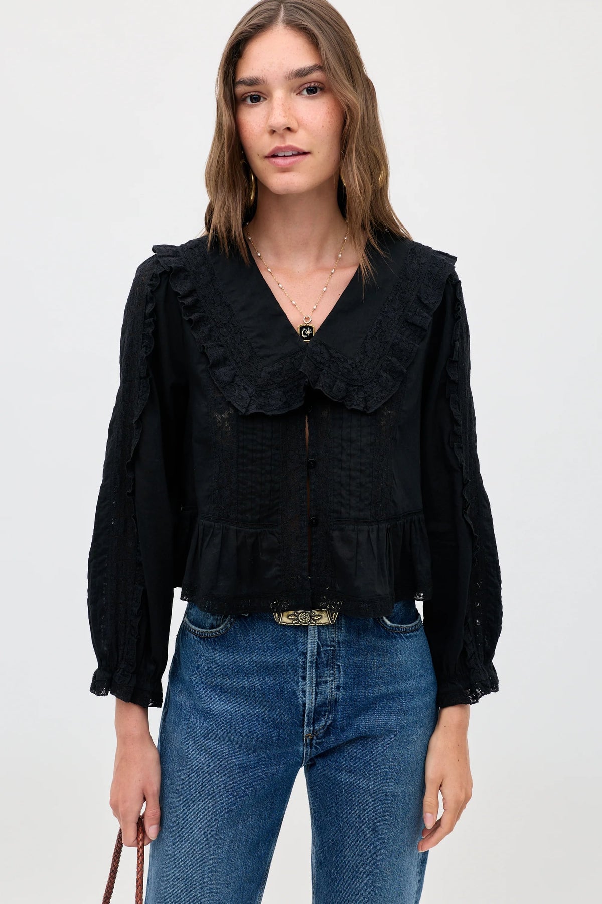 Woman with wavy brown hair wears the Maribelle - Black, a vintage-inspired blouse featuring long sleeves, ruffled details, a large collar, and a relaxed fit. She pairs it with flattering blue jeans and holds a small brown purse while standing against a plain light background.