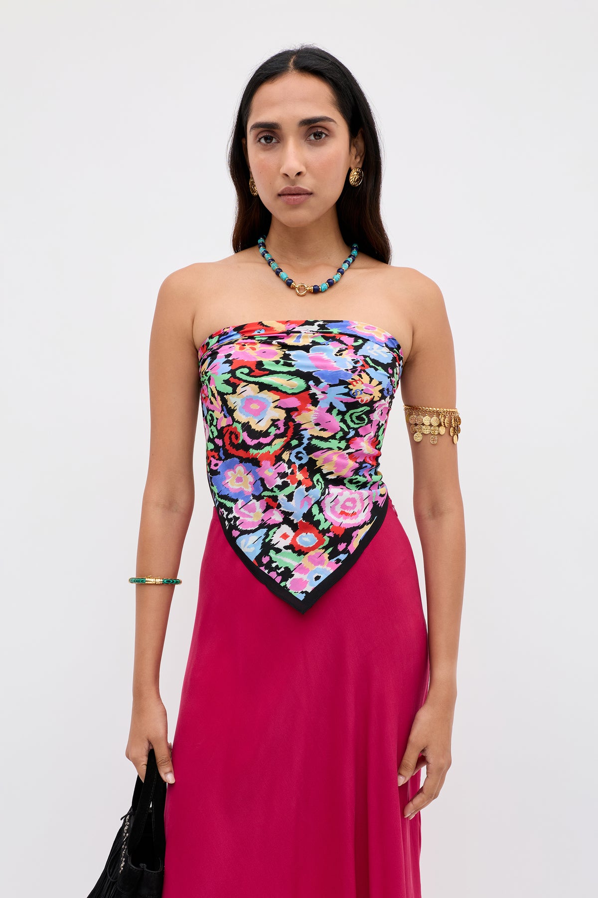 A woman stands against a plain white background, wearing the Menno - Ikat Bloom colorful floral scarf top, paired with a pink skirt, turquoise necklace, gold earrings, an arm bracelet, and holding a black bag.