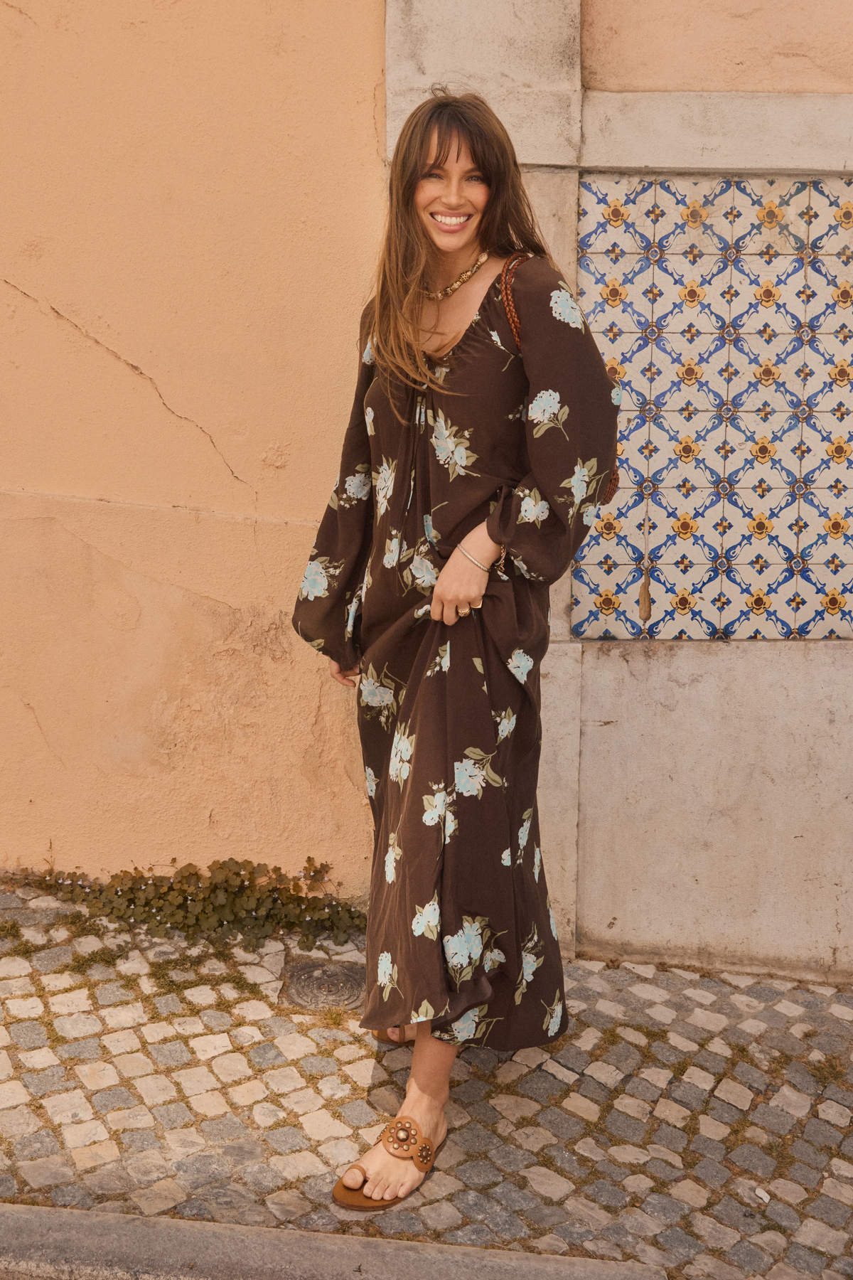 Wearing the Lianne - Peonies Scatter Brown dress, a woman smiles as she stands in sandals on a cobblestone street beside a peach-colored wall adorned with blue-and-white decorative tiles.