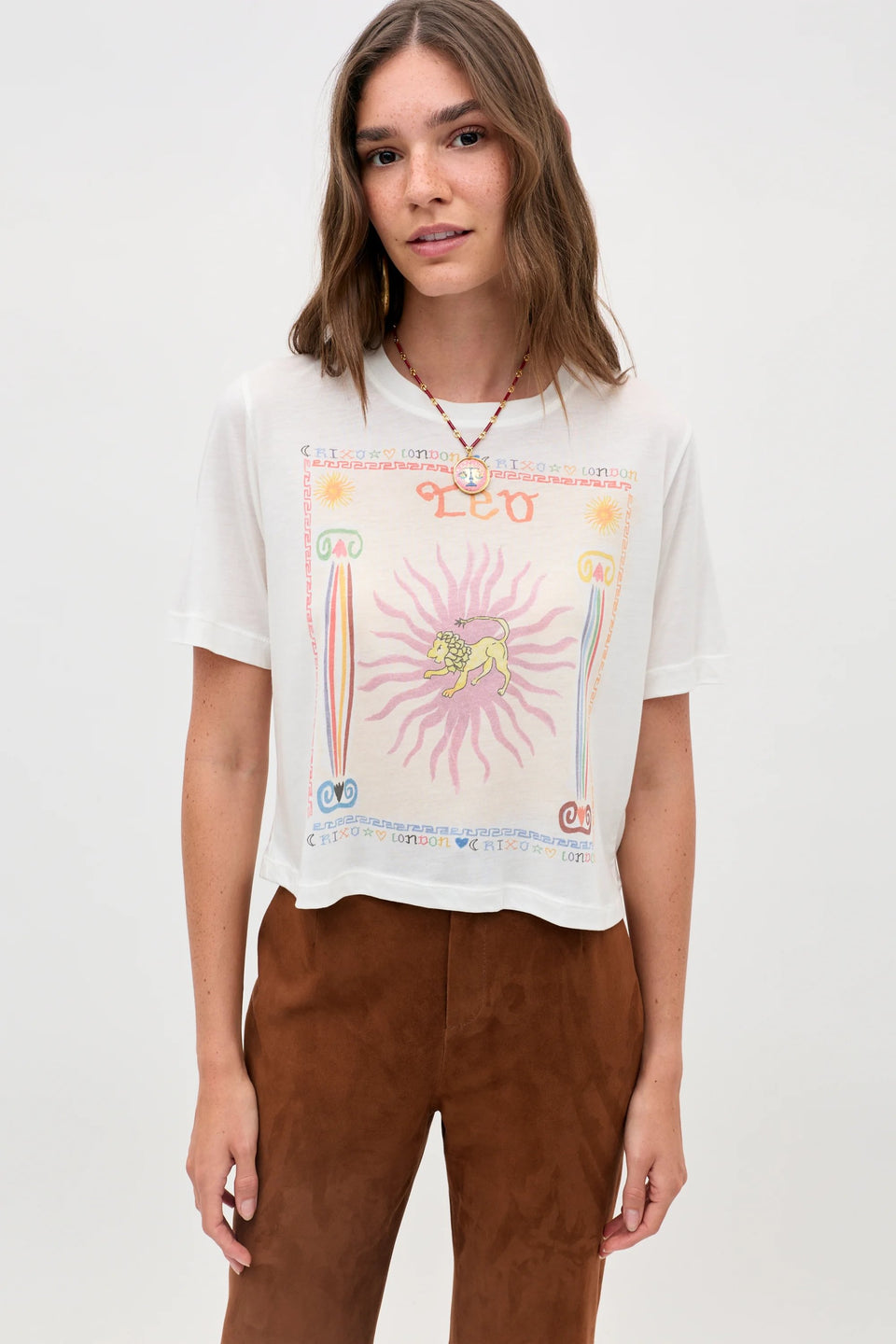 A woman with shoulder-length brown hair wears the Ria - Leo T-shirt, which features a vibrant, mystical Leo design, paired with brown pants. She stands against a plain light background, looking at the camera with a slight smile.