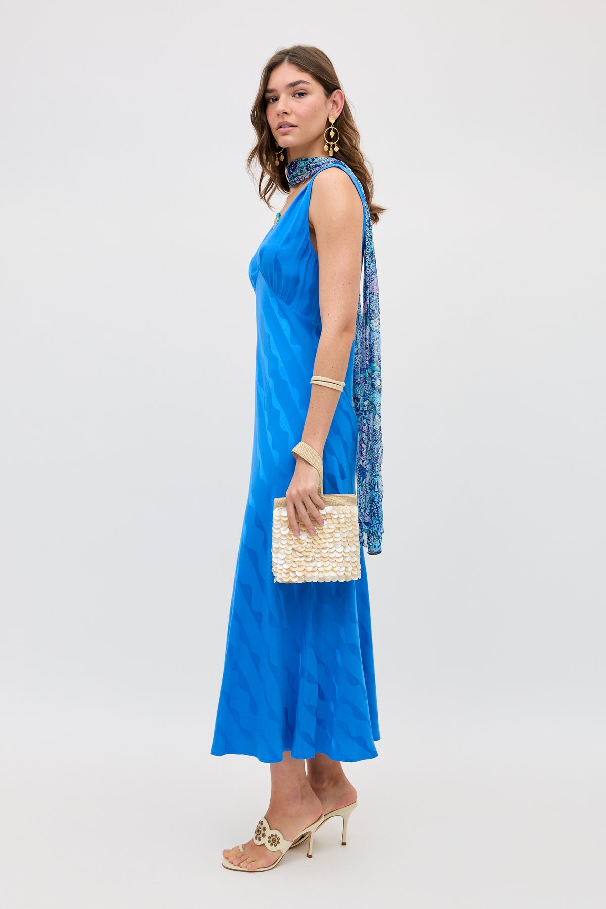 A woman in the Sandrine - Wave Jacquard Blue dress by RIXO, a standout transitional piece, styled with a patterned scarf draped over one shoulder, gold earrings and bracelet, heeled sandals, and a textured cream clutch.