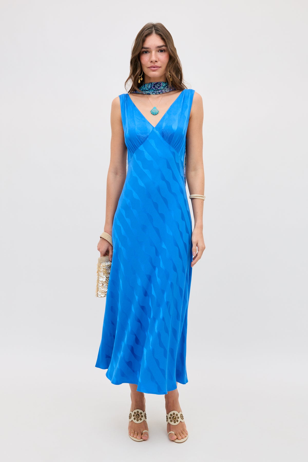 A woman wears the Sandrine - Wave Jacquard Blue dress, a sleeveless V-neck midi in bright blue with a wavy pattern, standing against a plain backdrop. She accessorizes with a gold clutch, heeled sandals, jewelry, and a choker necklace.
