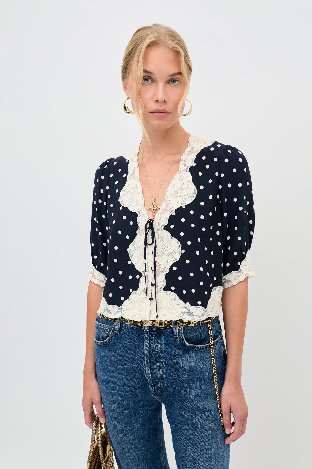 A woman wearing the ursula top layla dot navy blouse, paired with blue jeans, gold hoop earrings, and holding a gold chain bag, stands against a plain white background.