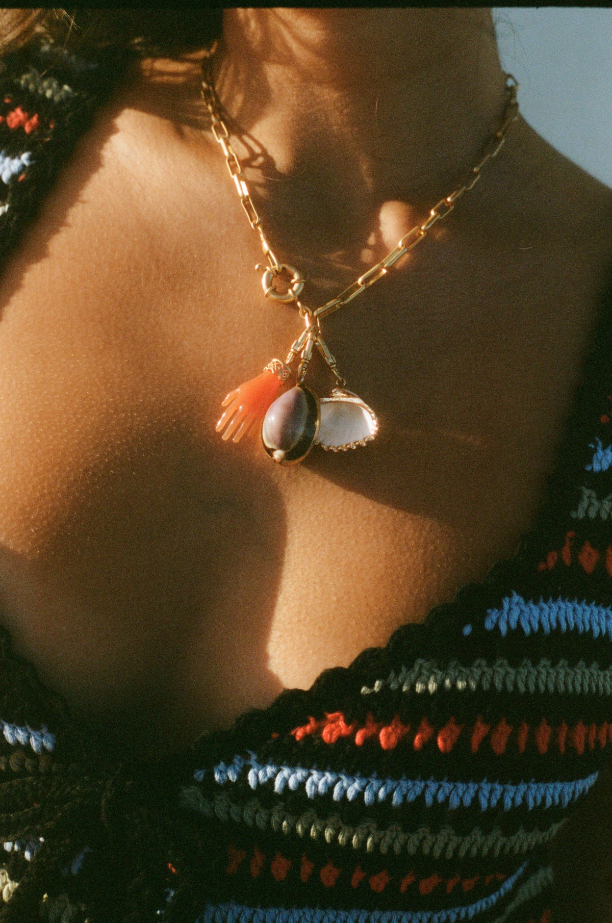 A close-up of a woman wearing the Hand - Orange necklace: a gold chain with an antique-style pendant featuring an orange hand charm, a purple stone, and a white shell. She pairs it with a blue, red, black, and white striped crocheted top.