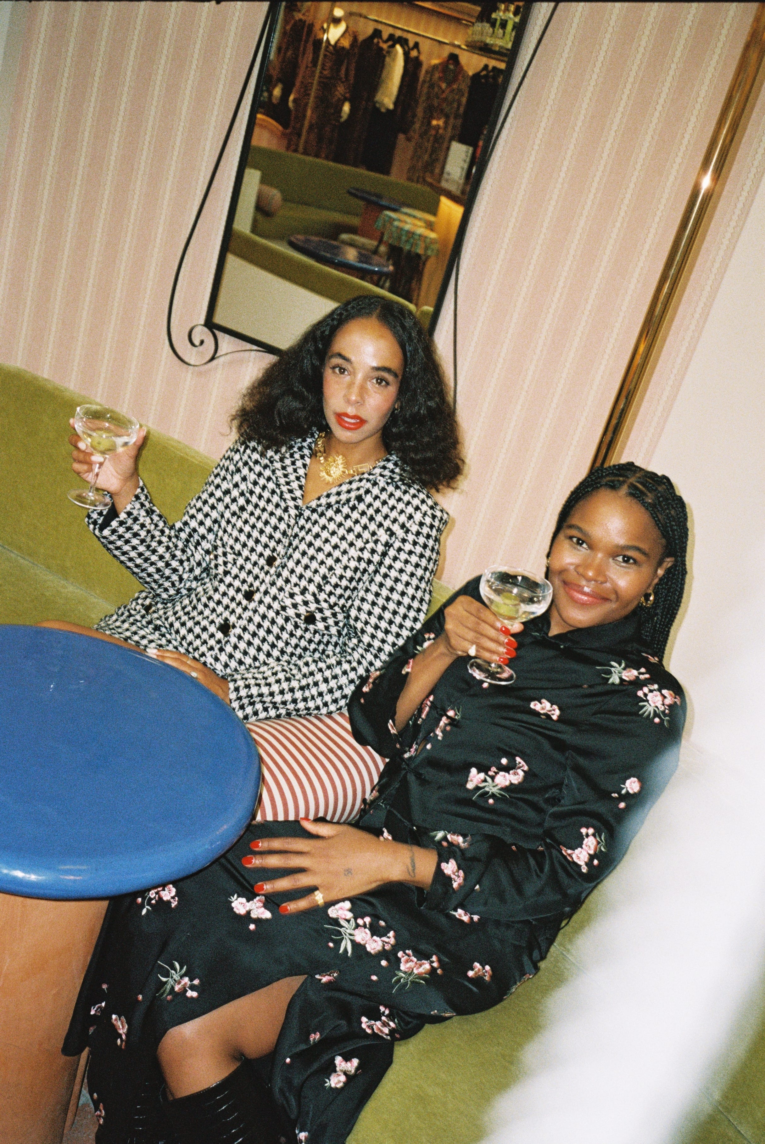 Two women sitting together, one holding a glass of wine, in a room with a mirror and clothing items.