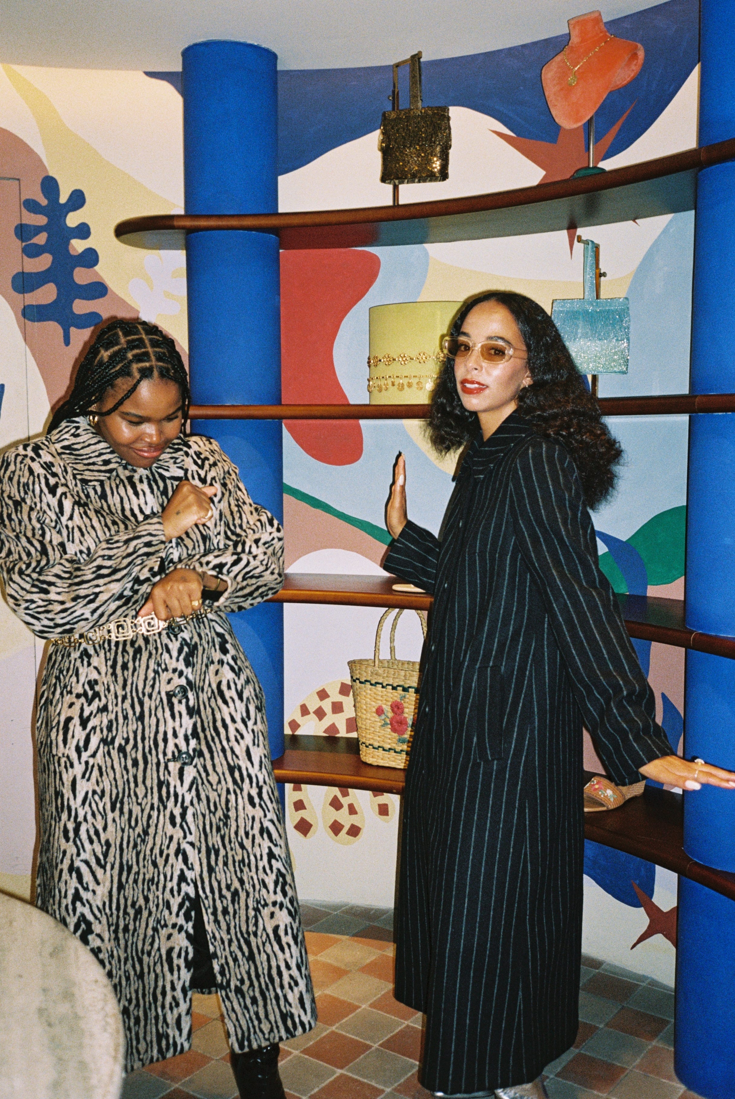Two women posing in front of a colorful wall with a shelf displaying bags.