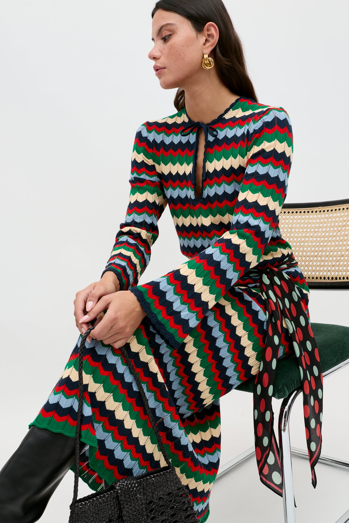 A woman sits on a chair wearing the Lucila - Retro Chevron Blue dress, which features a colorful zigzag pattern and a keyhole neckline. She pairs it with black boots, gold hoop earrings, and holds a black woven bag.