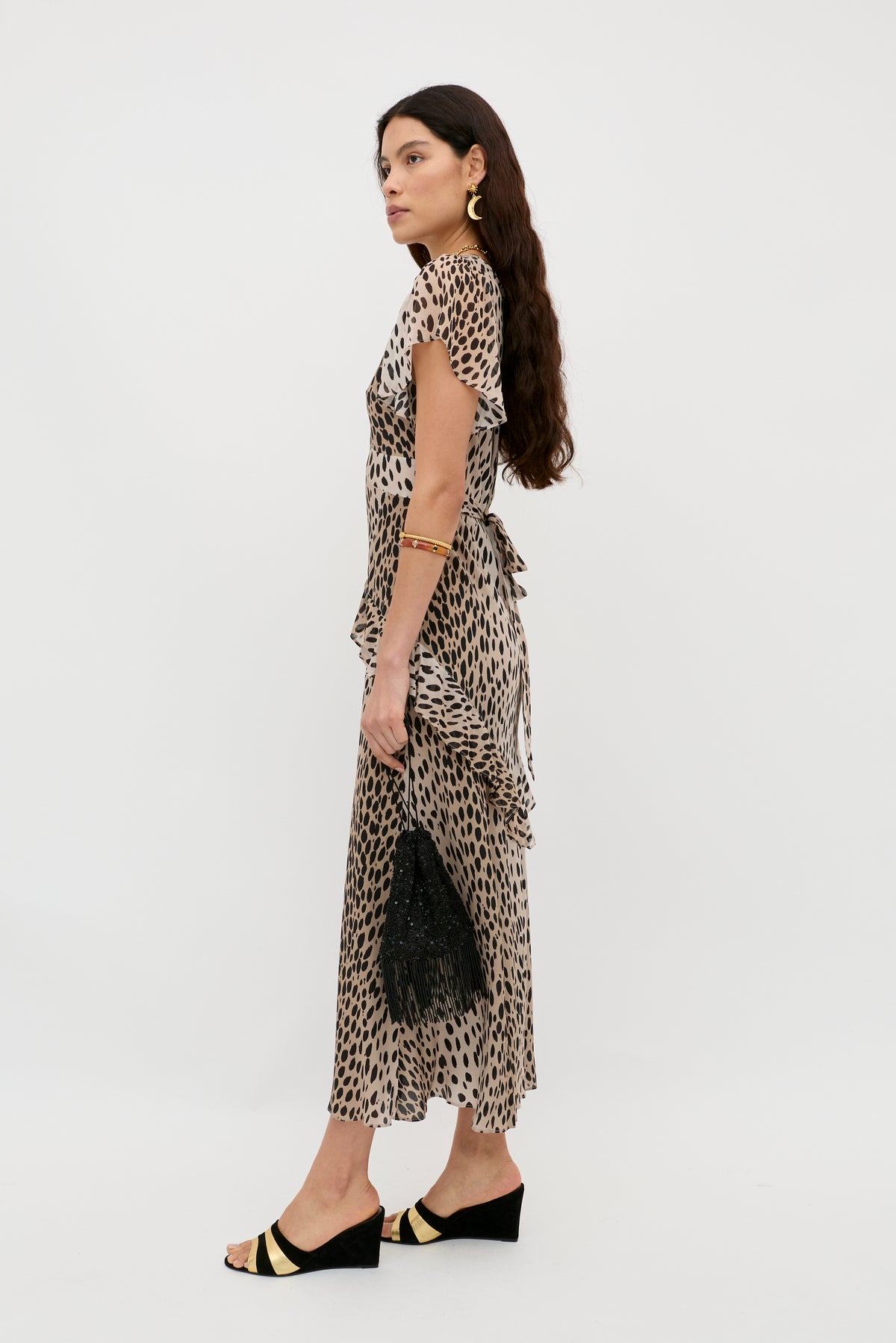A woman in profile wears the Evie - Antelope Spot Black ankle-length dress, black and gold wedge sandals, gold jewelry, and holds a black fringed bag. She has long wavy brown hair and a neutral expression.