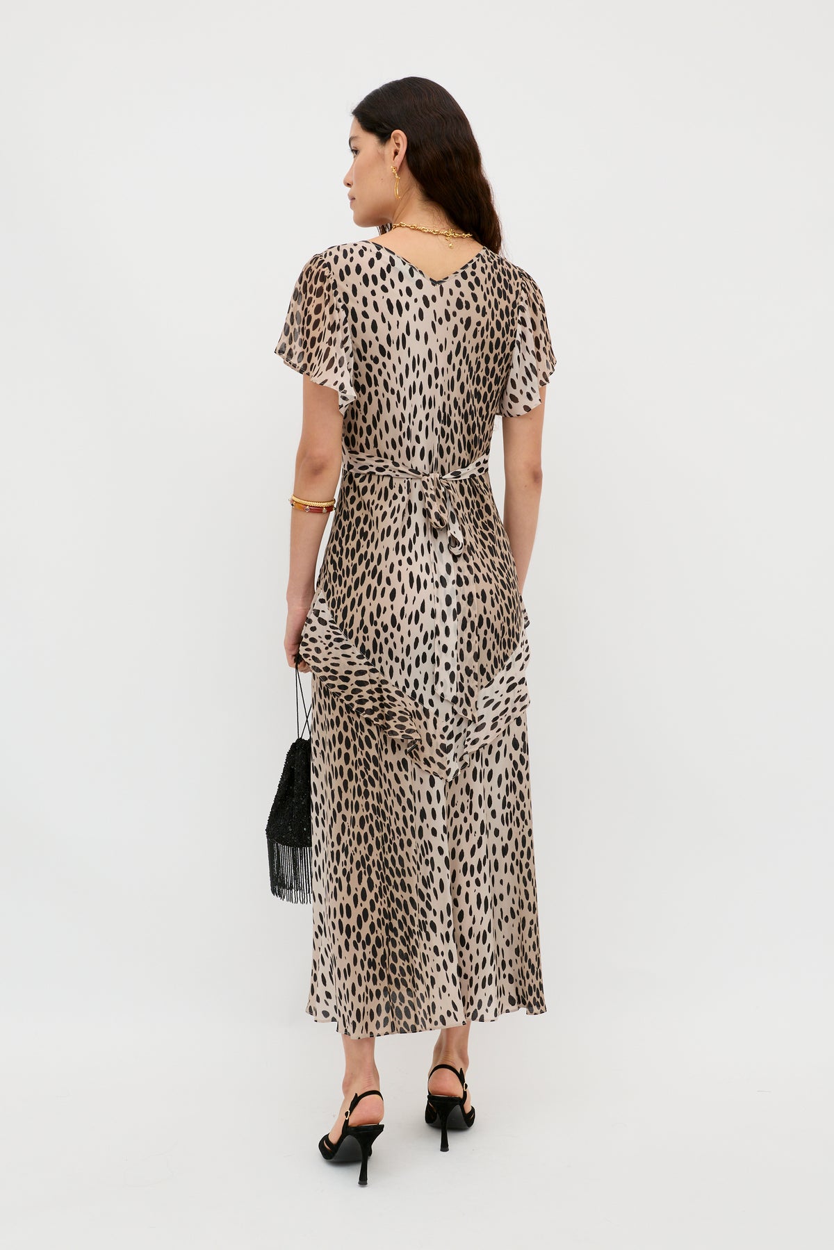 A woman faces away, wearing the mid-length, short-sleeved Evie - Antelope Spot Black dress with a tie belt. She carries a small black fringe bag and wears black heeled sandals, exuding bohemian style against a plain white background.
