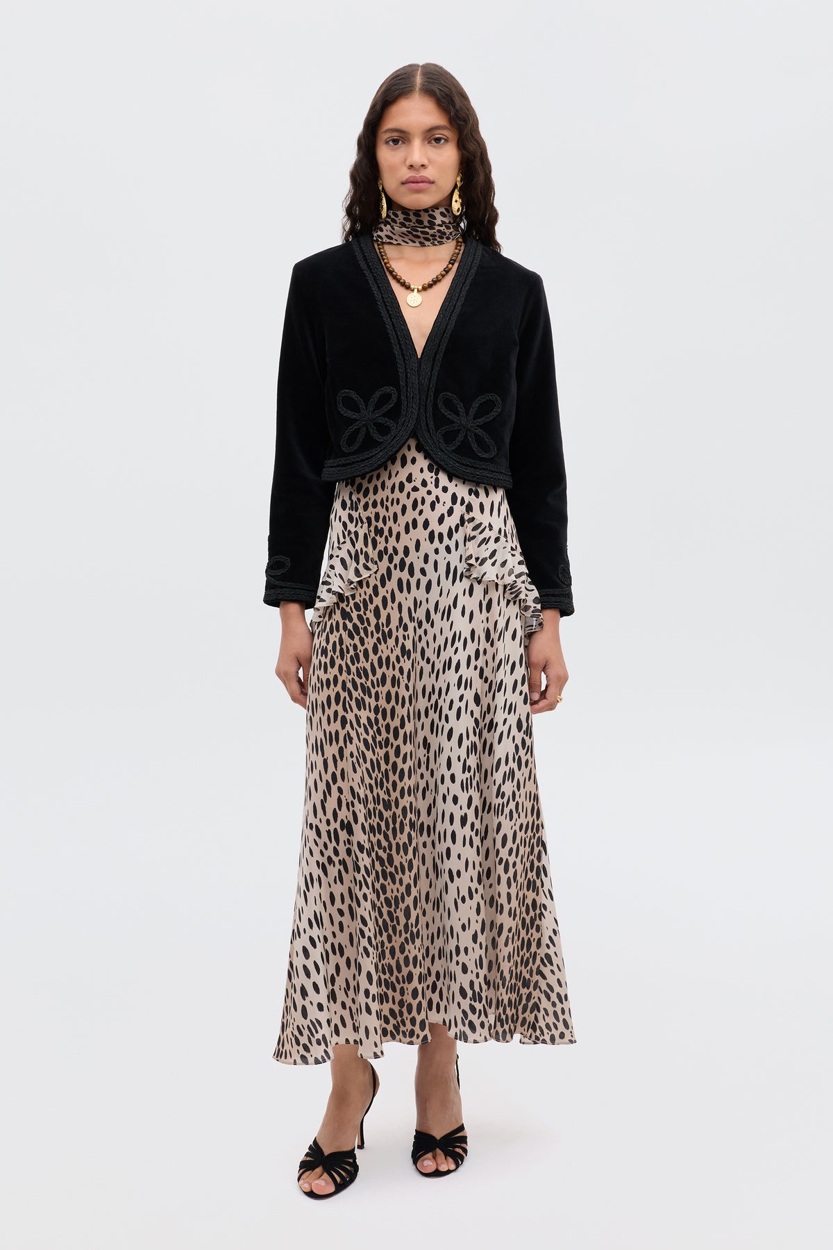 A woman with long, wavy brown hair wears the Evie - Antelope Spot Black skirt, styled with a black cropped jacket, layered necklaces, and black heeled sandals. She stands facing forward against a plain white background.