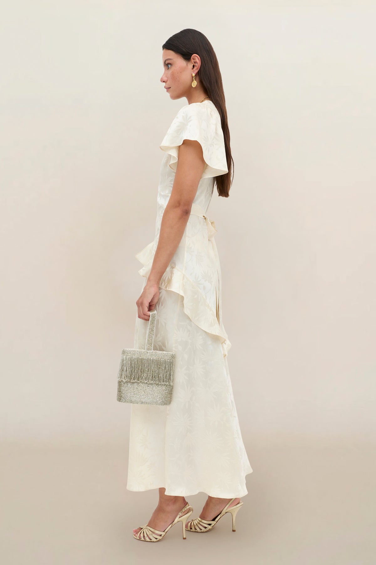 A woman stands sideways against a plain neutral background, wearing the Evie - Daisy Jacquard Champagne dress with elegant ruffles and heeled sandals. She holds a small gray fringed bag, accessorizing with gold earrings, and her straight brown hair completes the look.