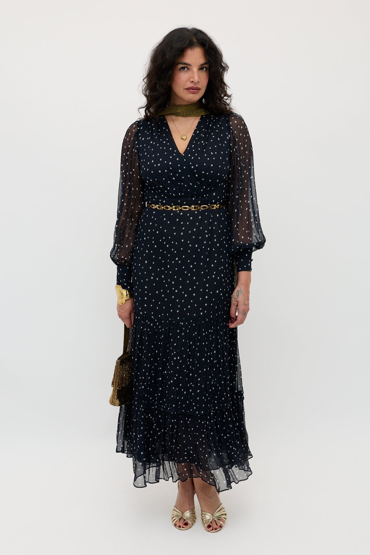 A woman stands against a plain background in the Elsie - Starry Night Navy dress—a long, sheer-sleeved navy dress with white dots—paired with gold accessories and open-toed shoes. Her wavy dark hair frames her neutral expression.
